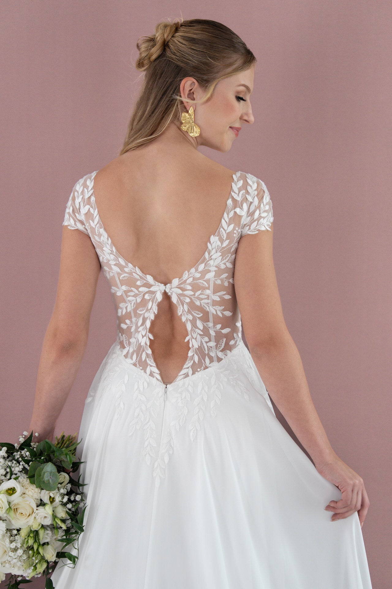 Eine Frau im weißen Brautkleid Bridonista V26-A05 mit durchsichtigem, blattgemustertem Rücken und Kappenärmeln hält einen Strauß weißer Blumen. Sie trägt goldene Ohrringe und ihr Haar ist zu einer Hochsteckfrisur frisiert, die vor einem zartrosa Hintergrund steht.