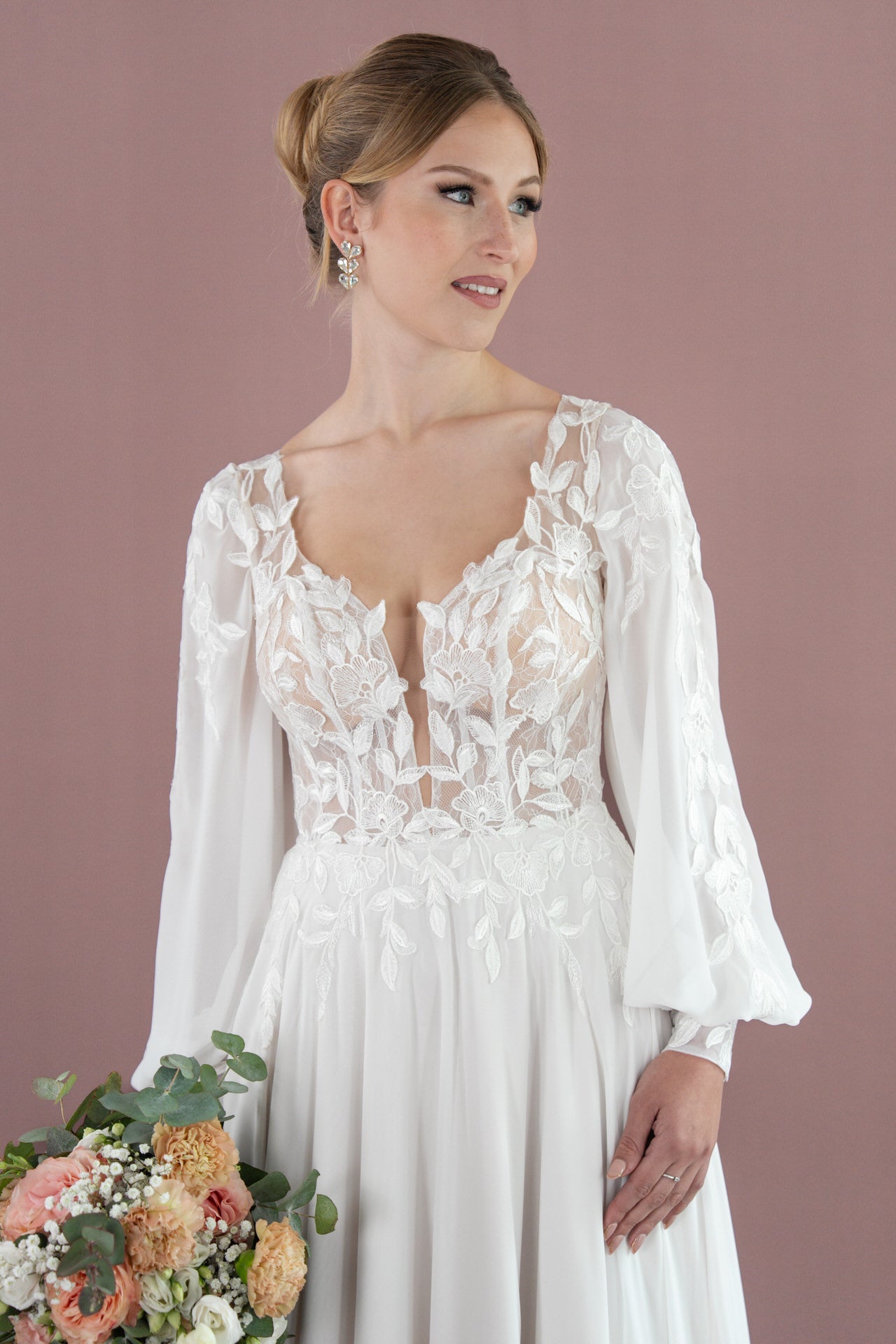 Eine Frau in Bridonista V26-A07, einem weißen, langärmeligen Brautkleid mit floralen Spitzendetails, hält einen Blumenstrauß und blickt zur Seite vor einem zartrosa Hintergrund.