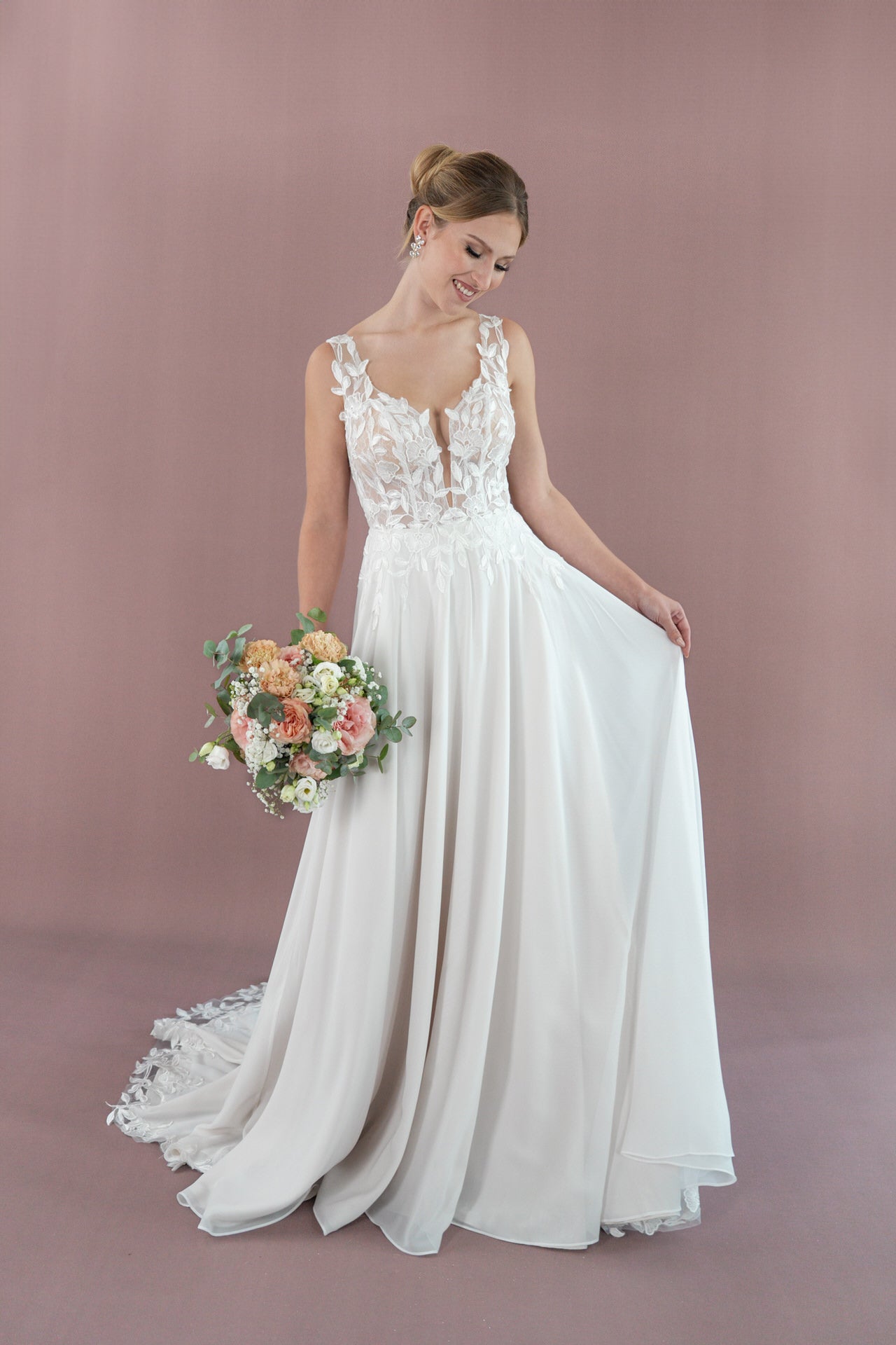 Eine Frau im Bridonista V26-A07, einem weißen, ärmellosen Brautkleid mit Blumendetails, hält lächelnd pastellfarbene Blumen vor einem hellvioletten Hintergrund.