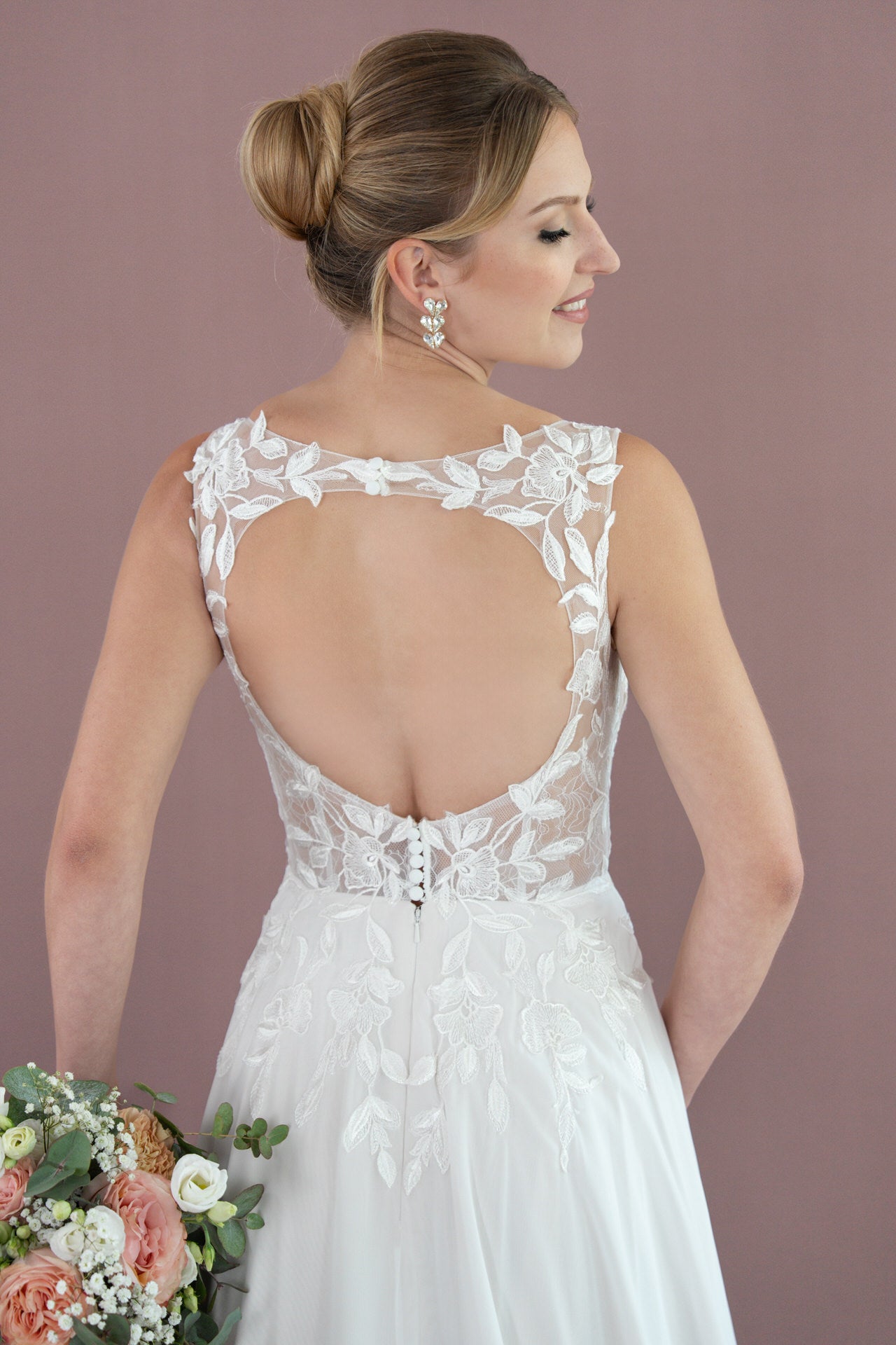Eine Braut im Bridonista V26-A07 Brautkleid aus weißer Spitze mit offenem Rücken und Blumendetails lächelt, hält einen Blumenstrauß in der Hand und trägt ihr Haar in einer eleganten Hochsteckfrisur vor einem zartlila Hintergrund.