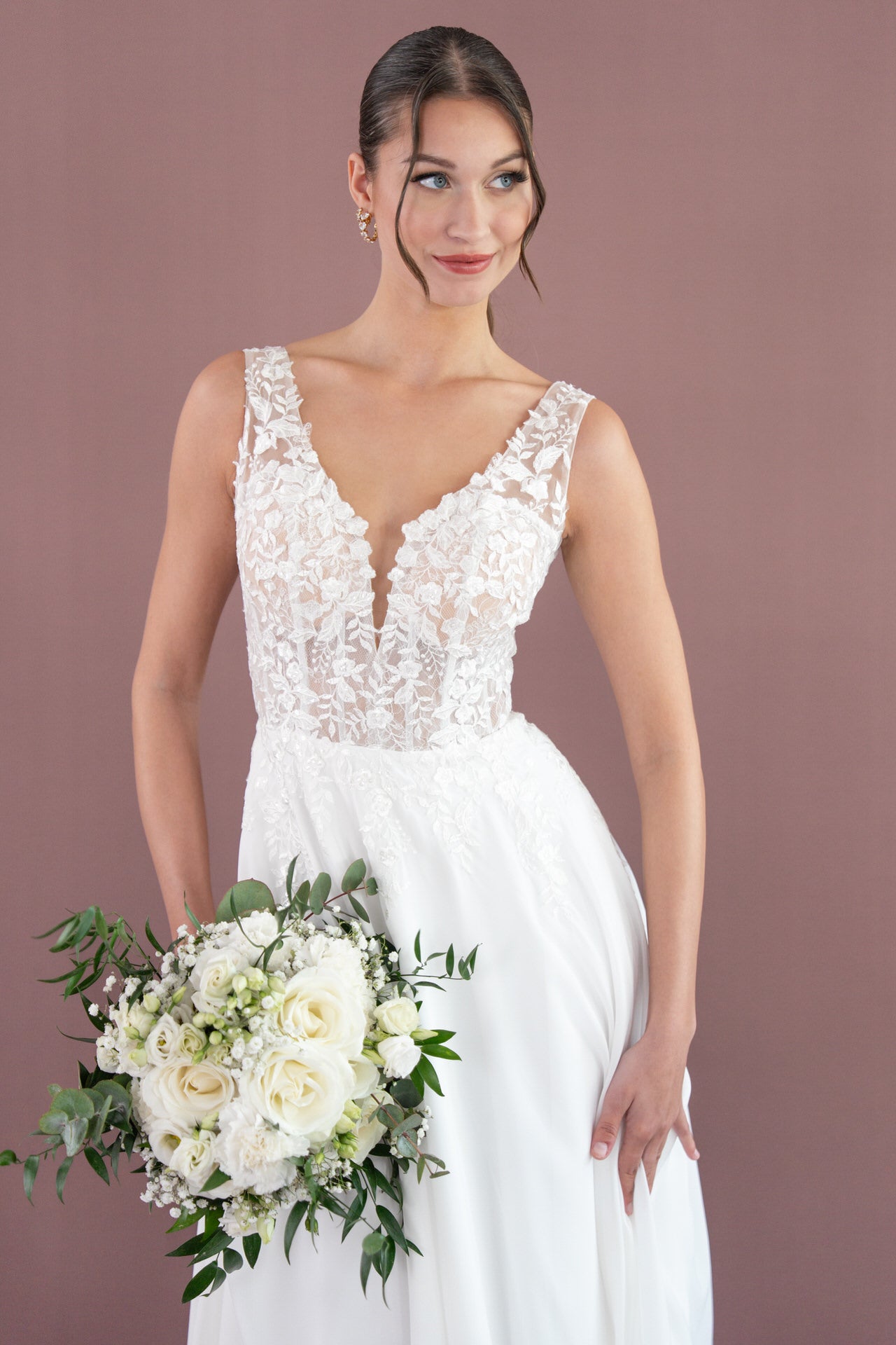 Eine Braut im ärmellosen Bridonista V26-A08 Brautkleid mit tiefem V-Ausschnitt hält einen Strauß weißer Rosen und Grünzeug, steht vor einem unifarbenen mauvefarbenen Hintergrund und lächelt sanft.