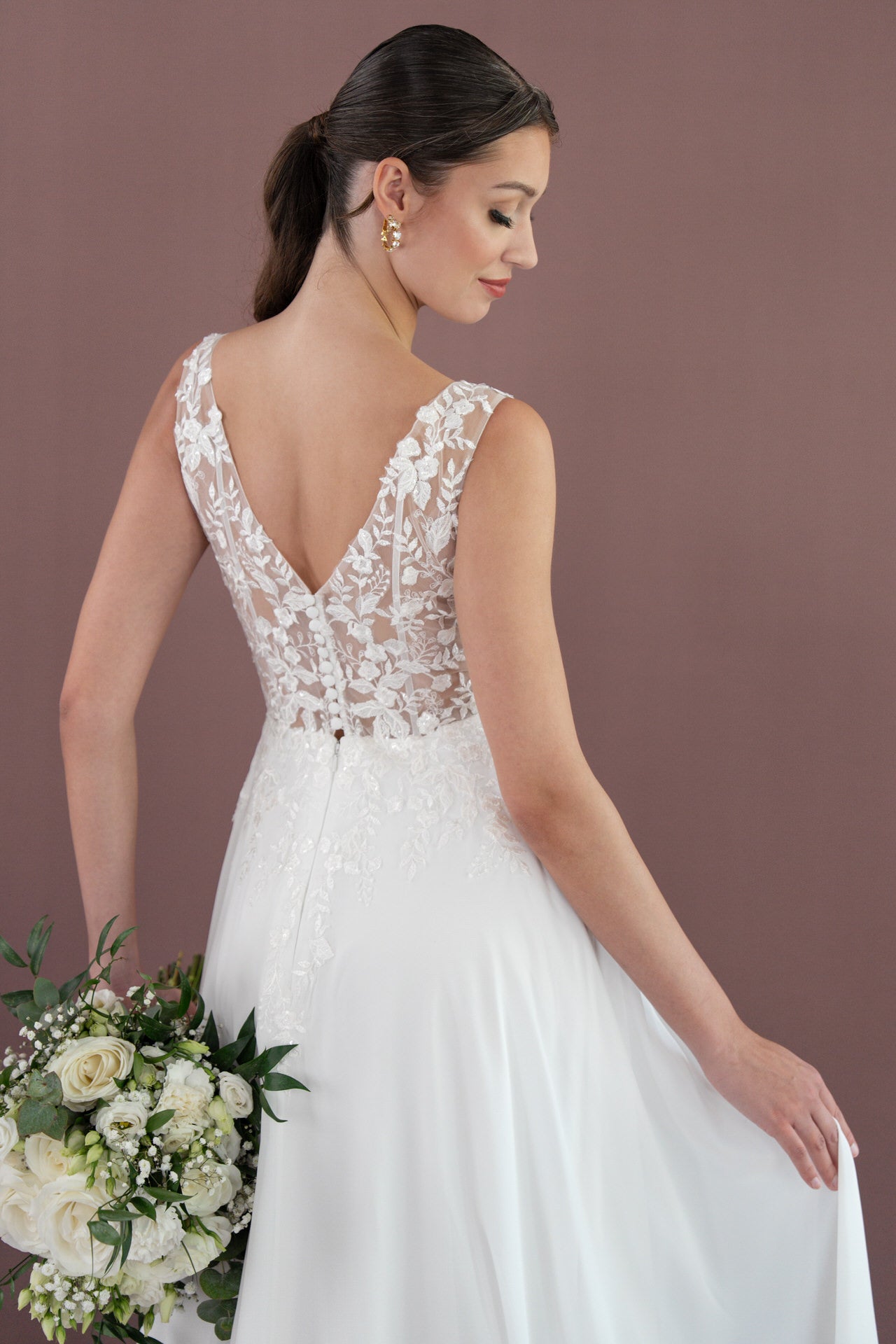 Eine Braut in dem ärmellosen Spitzenkleid Bridonista V26-A08 hält einen Strauß weißer Blumen und Grünzeug und wendet sich ab, um die aufwendigen Spitzen- und Knopfdetails zu präsentieren. Ihr Haar ist zu einem niedrigen Pferdeschwanz gebunden und steht vor einem schlichten Hintergrund.