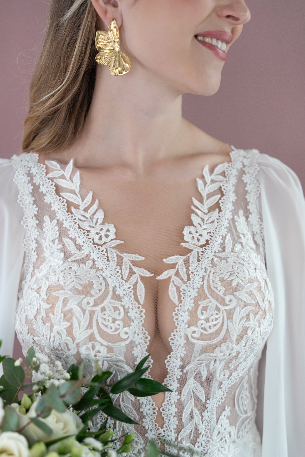 Eine Frau im Bridonista V26-A09 Brautkleid aus weißer Spitze mit durchsichtigen Ärmeln hält einen Blumenstrauß, trägt große Ohrringe in Form eines goldenen Blattes und lächelt vor einem rosa Hintergrund.