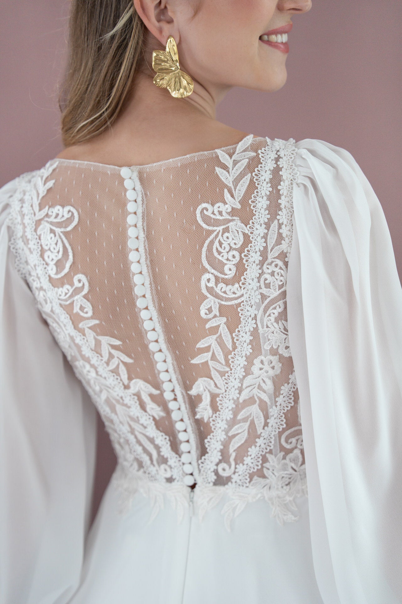 Eine Frau trägt das Bridonista V26-A09, ein weißes Brautkleid mit durchsichtiger Spitze und stoffbezogenen Knöpfen am Rücken. Sie kombiniert es mit goldenen Schmetterlingsohrringen und lächelt, von der Seite auf einem zartrosa Hintergrund gezeigt - eine ideale Brautmoden-Inspiration.