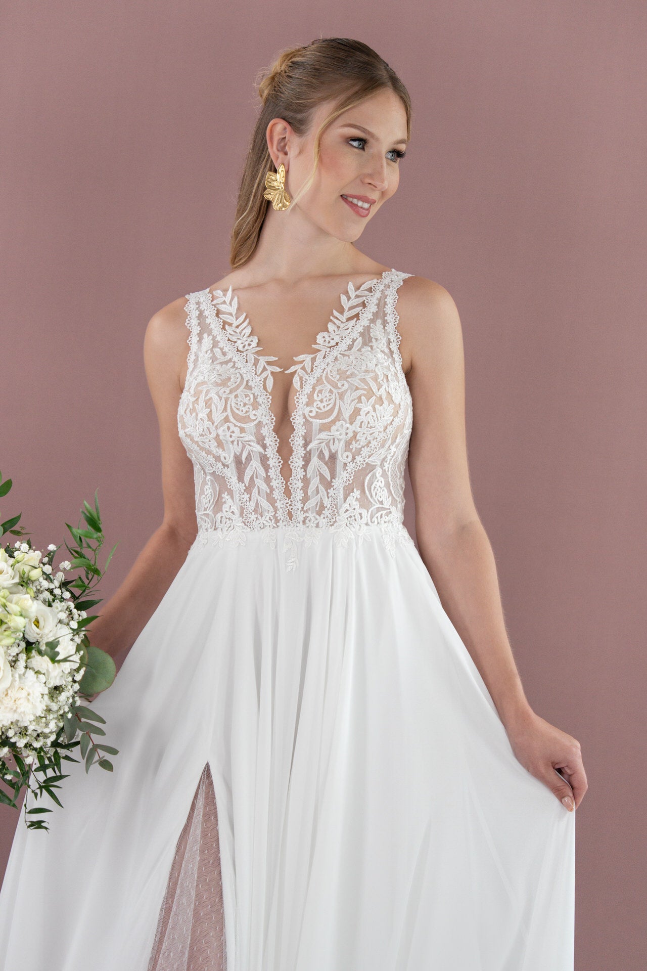 Eine Frau in dem weißen Spitzenbrautkleid Bridonista V26-A09 mit durchsichtigem, besticktem Mieder und einem fließenden Rock mit Schlitz hält weiße Blumen. Ihr hellbraunes Haar ist zurückgestylt, während sie vor einem malvenfarbenen Hintergrund posiert.