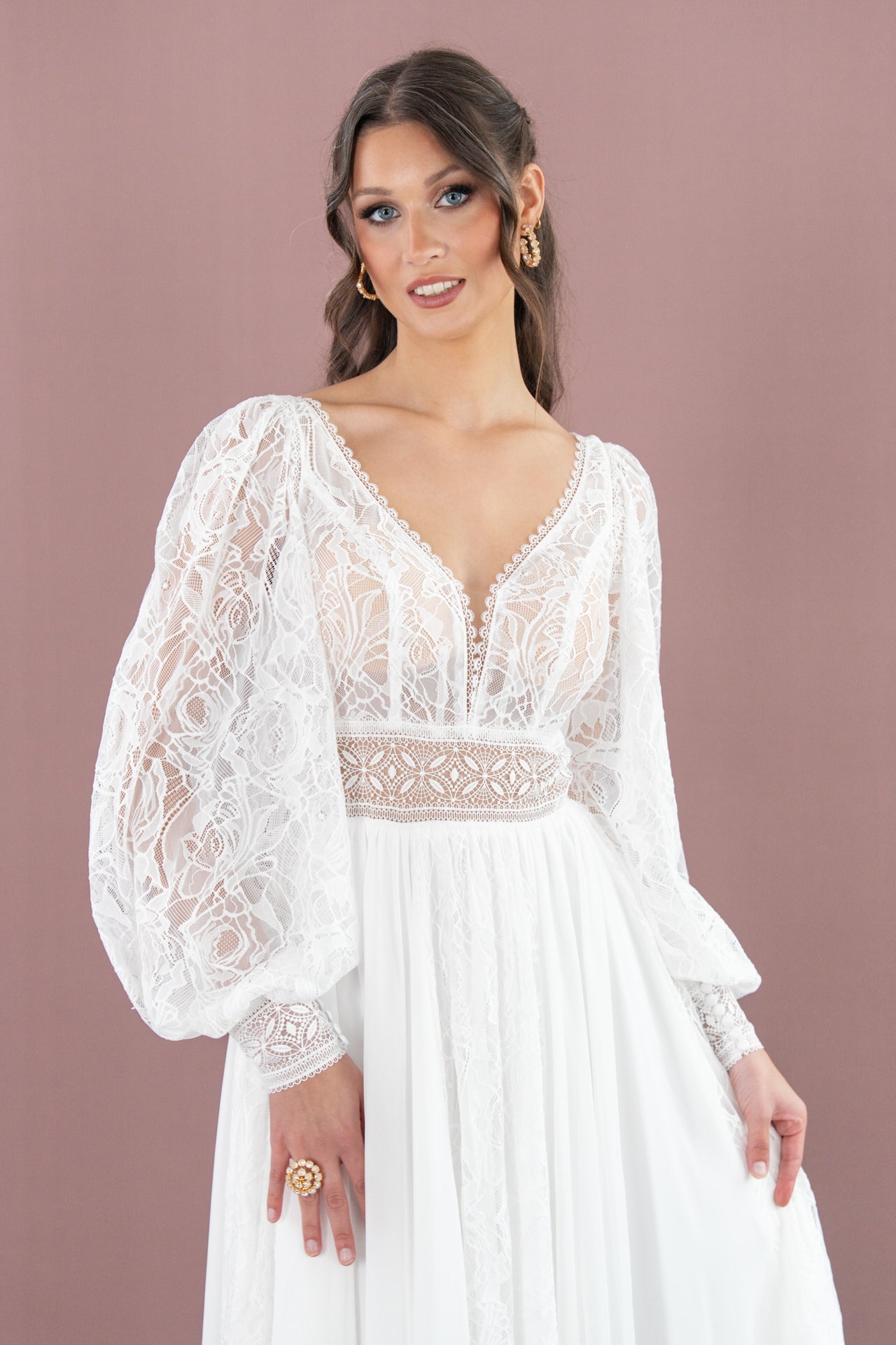Eine Frau im Bridonista V26-A10 Brautkleid aus weißer Spitze mit Puffärmeln, tiefem V-Ausschnitt und aufwändiger Stickerei steht lächelnd vor einem lilafarbenen Hintergrund und trägt ihr Haar in lockeren Wellen.