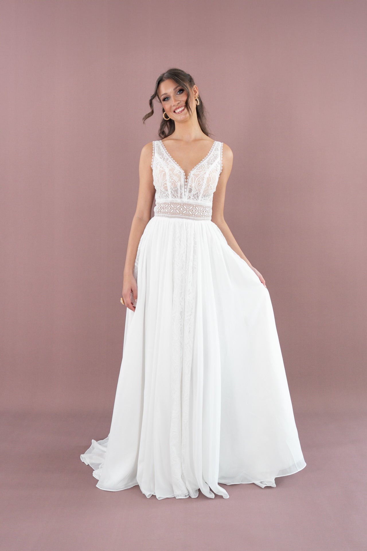 Eine Frau trägt das Brautkleid Bridonista V26-A10 mit Spitzencorsage und fließendem Rock, steht vor einem mauvefarbenen Hintergrund, lächelt und hält einen Teil ihres Kleides zur Seite.