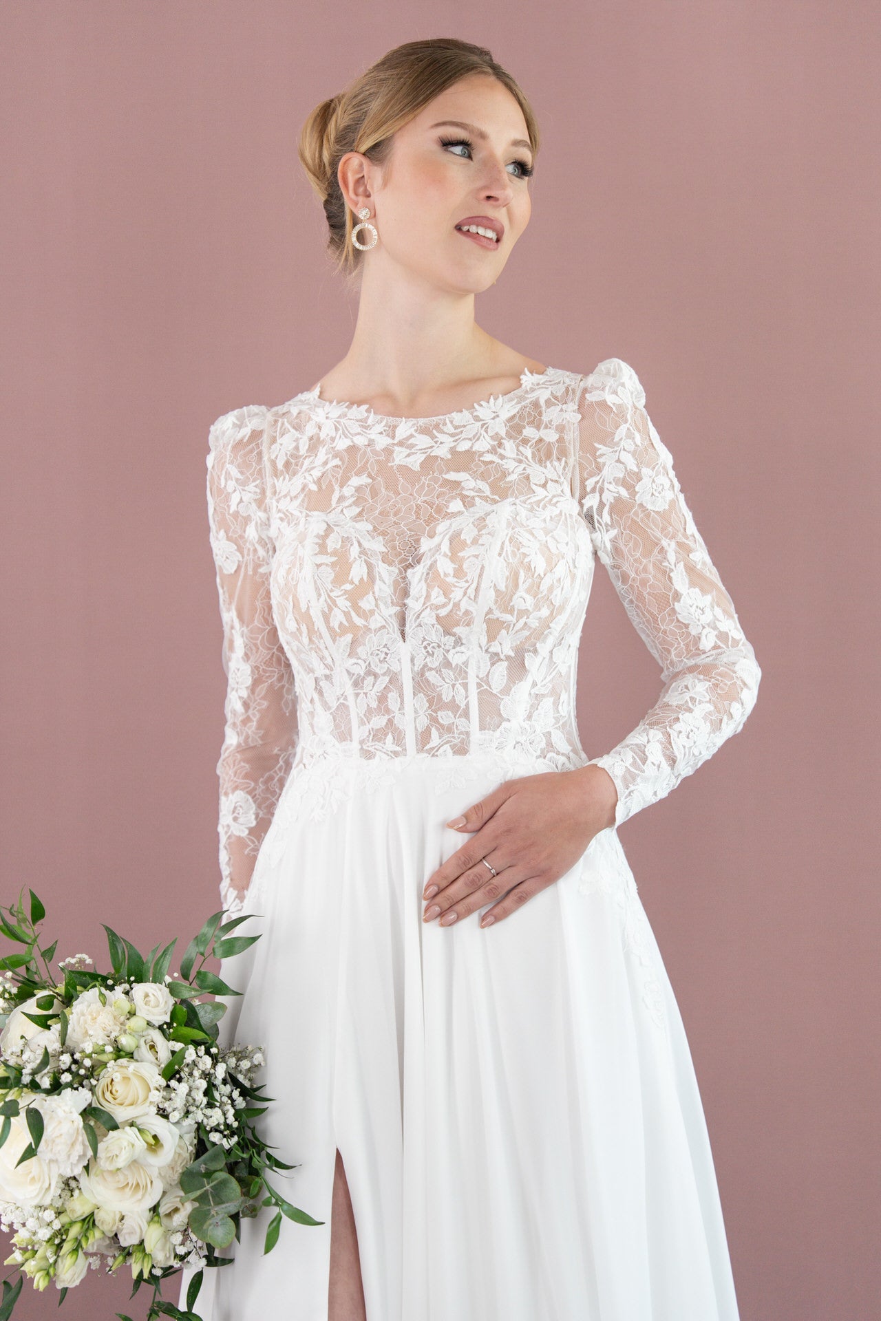Eine Frau im Bridonista V26-A12 Brautkleid aus weißer Spitze hält einen Strauß weißer Blumen und Grünzeug und steht vor einem malvenfarbenen Hintergrund. Ihr Haar ist zu einer Hochsteckfrisur frisiert und sie trägt Tropfenohrringe.