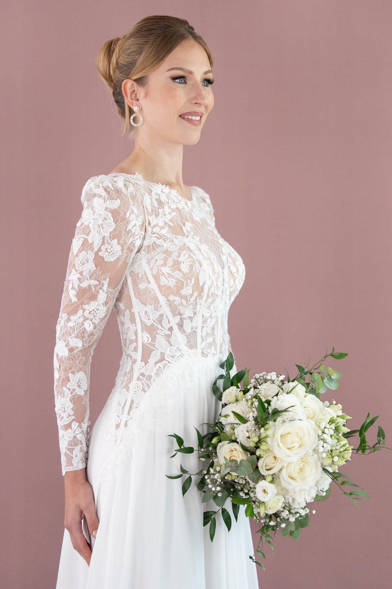 Eine Braut im weißen Spitzenhochzeitskleid V26-A12 von Bridonista steht vor einem lilafarbenen Hintergrund und hält einen Strauß aus weißen Rosen und Grünzeug. Ihr Haar ist zu einer Hochsteckfrisur gestylt, und sie lächelt, während sie leicht nach links blickt.