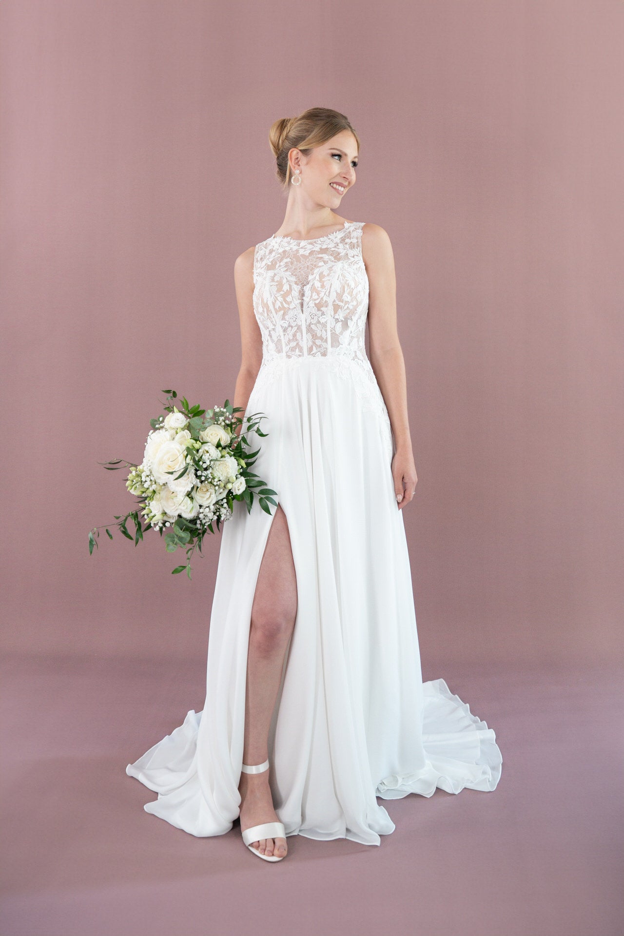 Eine Frau in Bridonista V26-A12, einem weißen ärmellosen Brautkleid mit Spitzenmieder und hohem Schlitz, hält einen Strauß weißer Blumen und Grünzeug vor einem zartrosa Hintergrund.