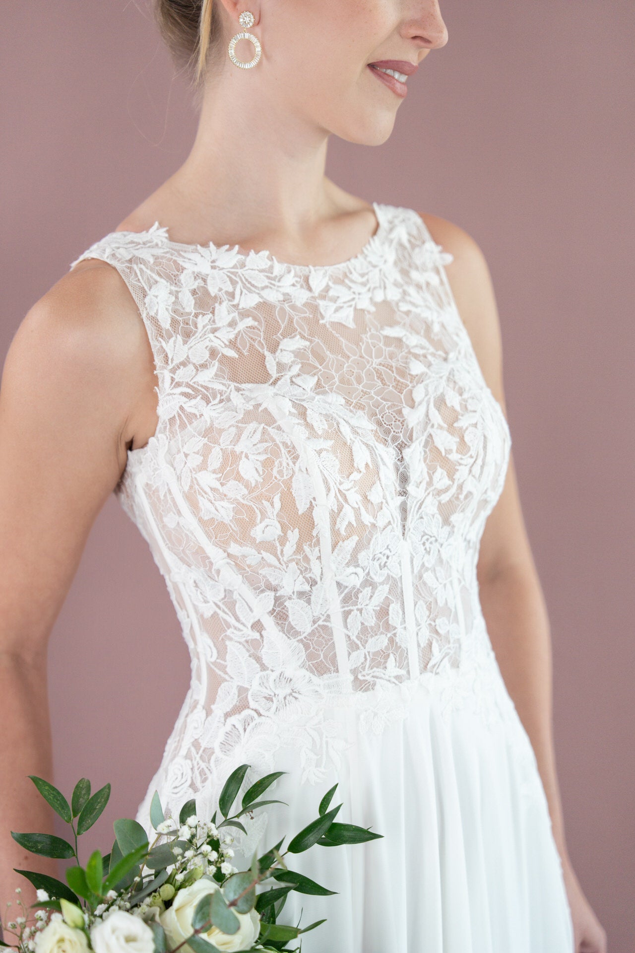 Eine Braut modelliert das Bridonista V26-A12, ein ärmelloses Brautkleid aus Spitze mit Blumenstickerei, und hält einen Strauß aus weißen Blüten und Grünzeug vor einem zartrosa Hintergrund. Der Oberkörper und ein Teil des Gesichts sind sichtbar - ideal für Brautmoden-Inspiration.
