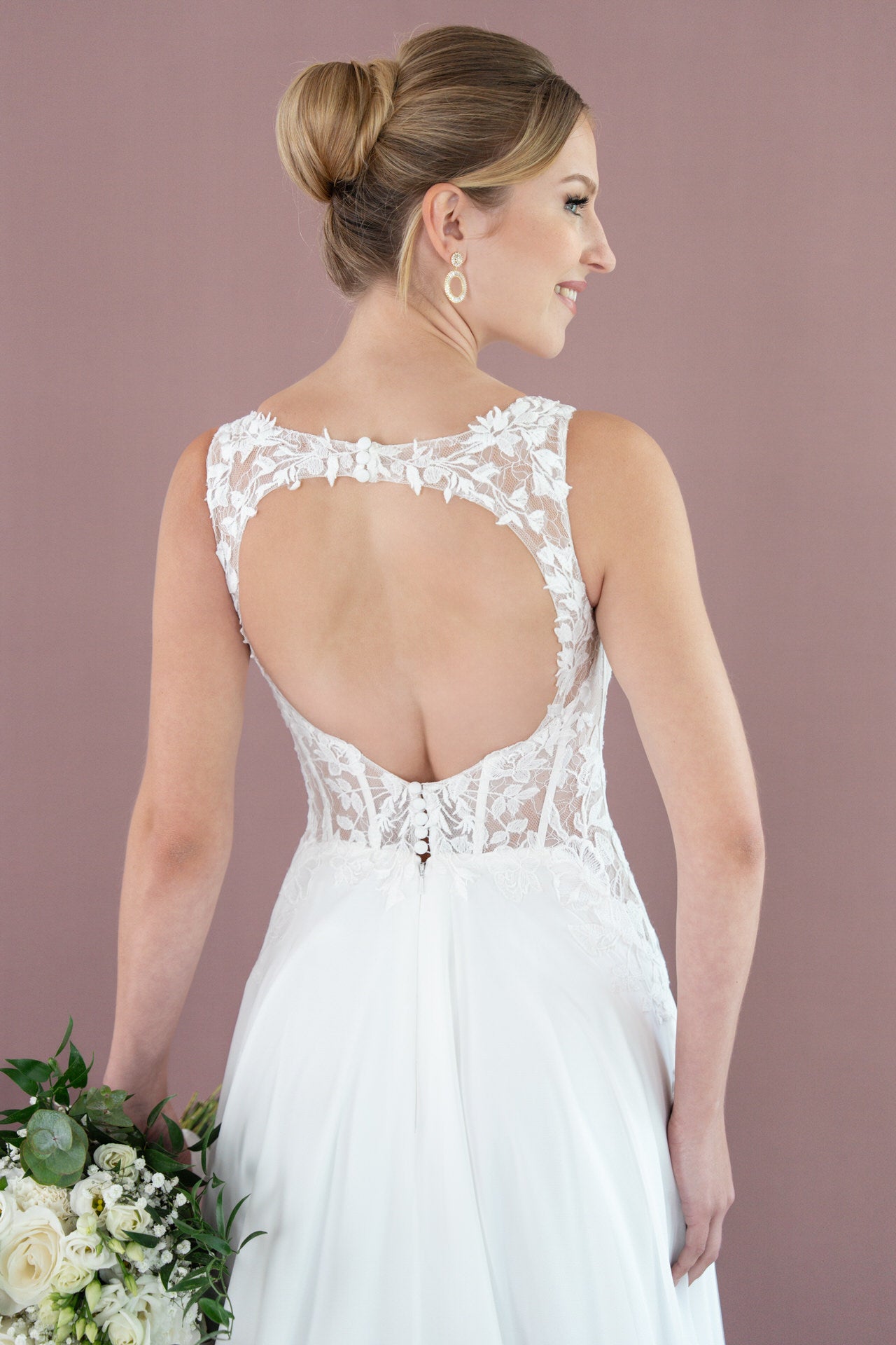 Eine Frau trägt das elegante Bridonista V26-A12 Brautkleid mit Spitze und offenem Rücken. Sie steht mit einem weißen Blumen-Bouquet vor mauvefarbenem Hintergrund, ihr Haar ist zu einem ordentlichen Dutt gestylt.