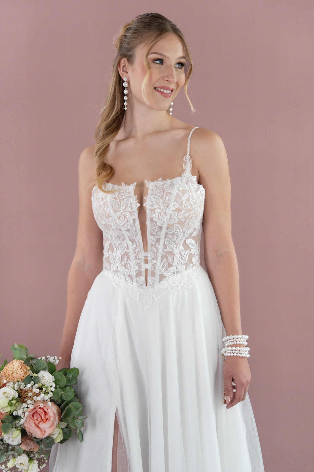 Eine Frau in einem weißen Bridonista V26-A14 Brautkleid hält einen Blumenstrauß in der Hand und lächelt, während sie vor einem malvenfarbenen Hintergrund steht. Sie hat langes, gewelltes blondes Haar, trägt baumelnde Ohrringe und Perlenarmbänder und schaut zur Seite.