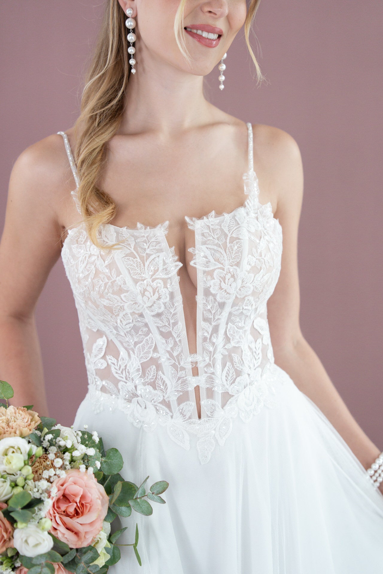 Eine Frau trägt das Bridonista V26-A14, ein weißes Spitzenbrautkleid mit einem durchsichtigen, blumenbestickten Mieder. Sie hält einen Blumenstrauß, hat gewelltes blondes Haar und Perlenohrringe und lächelt vor einem malvenfarbenen Hintergrund.