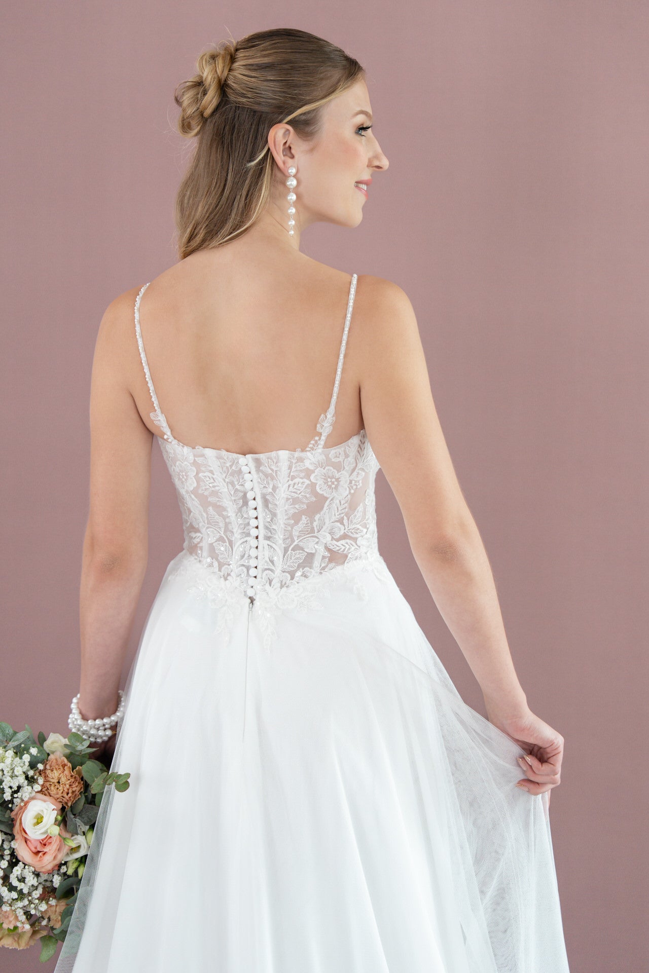 Eine Braut in dem weißen Spitzenkleid V26-A14 von Bridonista mit dünnen Trägern steht vor einem lilafarbenen Hintergrund und hält einen Blumenstrauß. Ihr Haar ist zu einer eleganten Hochsteckfrisur frisiert, die durch lange Perlenohrringe ergänzt wird.