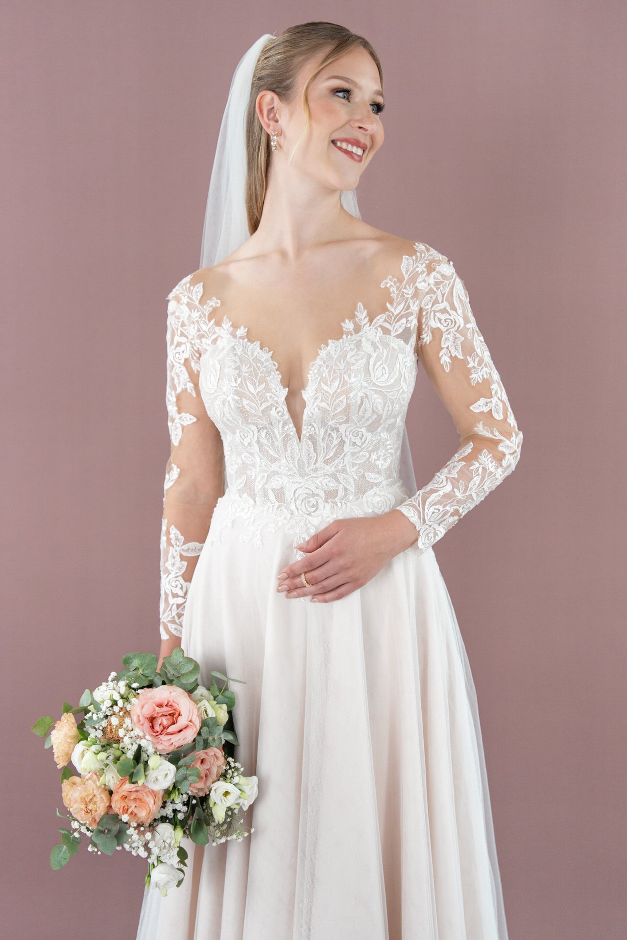 Eine lächelnde Braut im Bridonista V26-A17 Brautkleid aus weißer Spitze mit langen Ärmeln hält einen Strauß aus rosa, pfirsichfarbenen und weißen Blumen. Sie trägt einen Schleier und steht vor einem mauvefarbenen Hintergrund, der stilvolle Brautmoden hervorhebt.