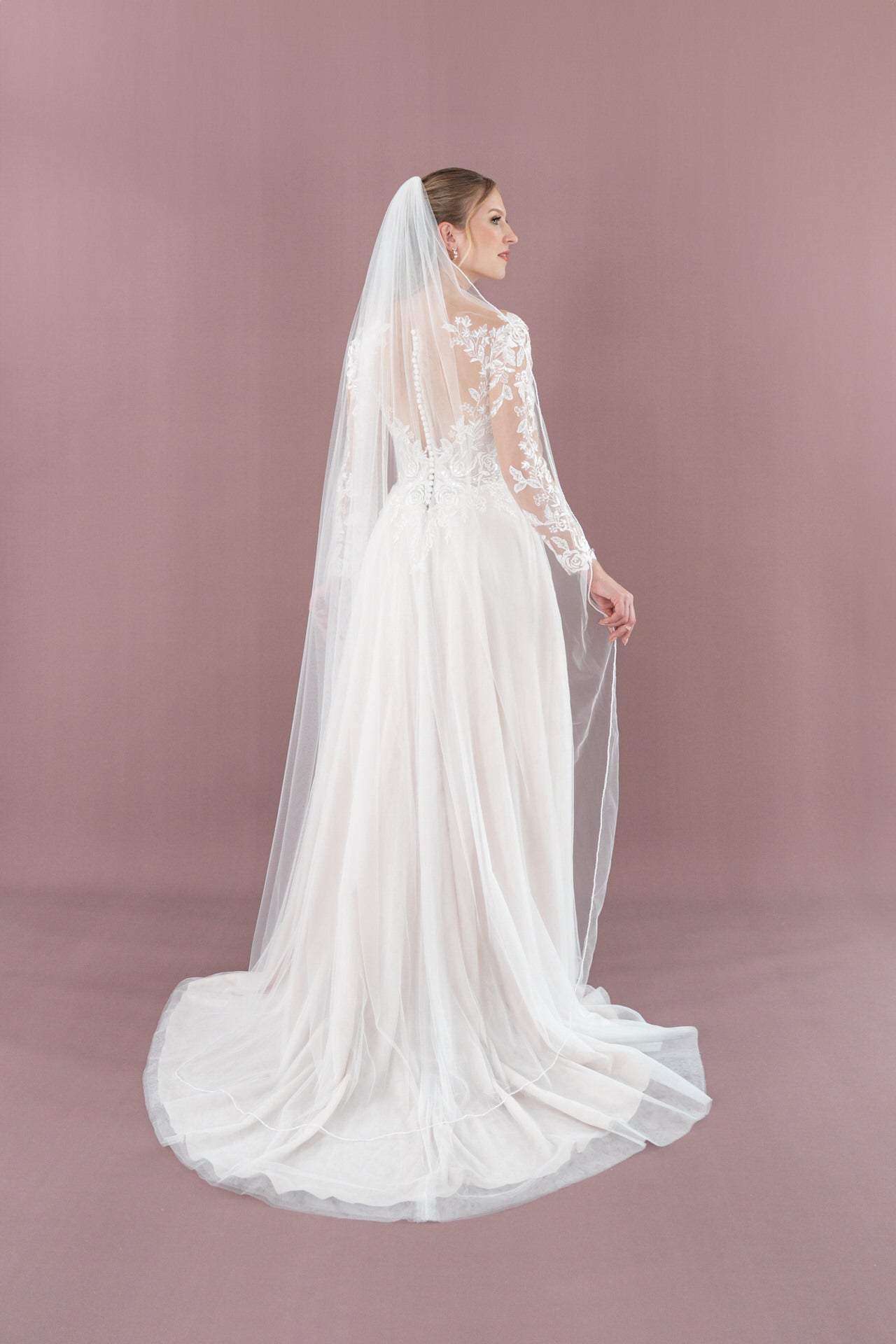 Eine Braut blickt von sich weg und trägt das Bridonista V26-A17 - ein langes weißes Kleid mit Spitzenärmeln und einem durchsichtigen bodenlangen Schleier - vor einem einfarbig lilafarbenen Hintergrund.