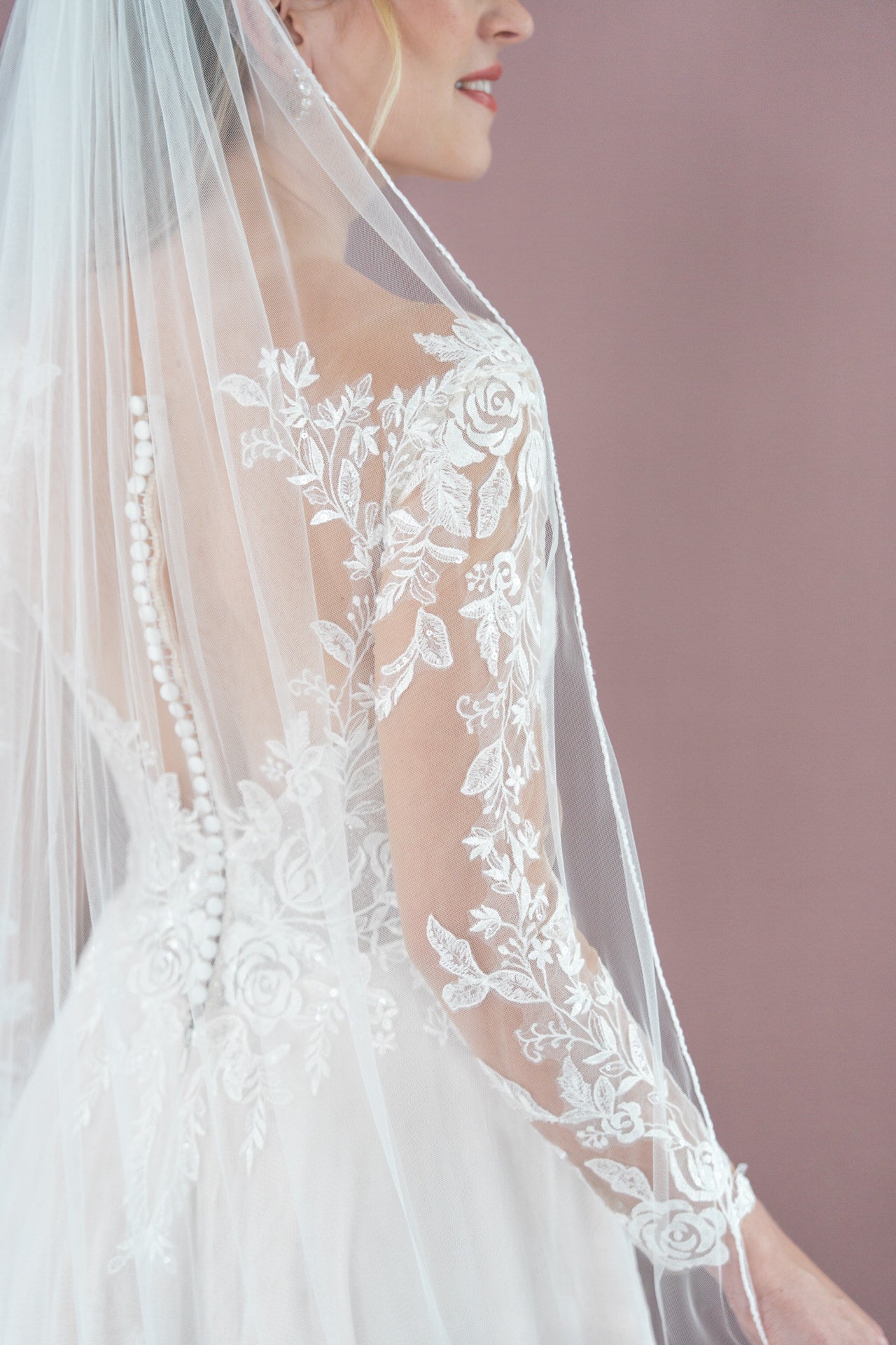 Eine Braut trägt das Bridonista V26-A17, ein weißes Spitzenbrautkleid mit floraler Stickerei und durchsichtigem Schleier, der aufwendige Rückendetails und Knöpfe vor einem zartrosa Hintergrund hervorhebt.