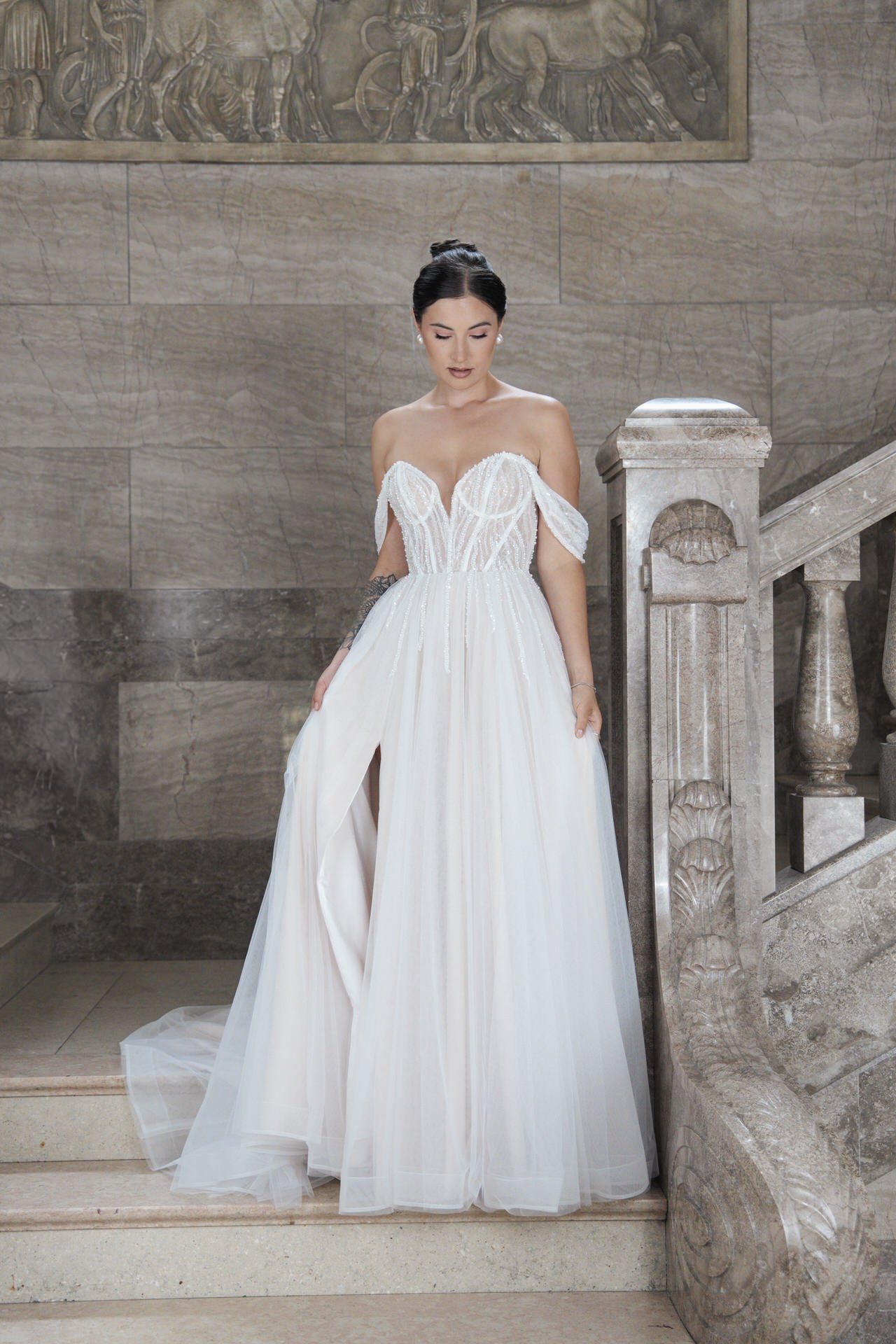 Eine Frau trägt das elegante Bridonista V26-A18: ein schulterfreies weißes Brautkleid mit transparenter Korsage und fließendem Rock. Auf Marmortreppen mit kunstvollem Steinwerk-Inspiration für moderne Brautmode.