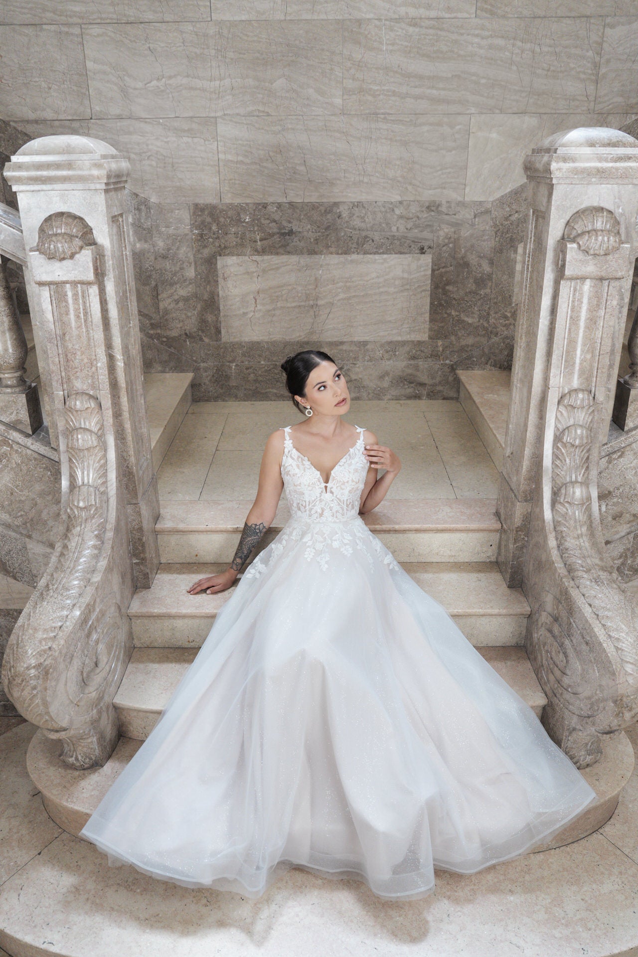Eine Frau trägt das elegante, weiße Hochzeitskleid Bridonista V26-A22, sitzt auf Marmortreppen und blickt nach oben. Kunstvolle Steingeländer und eine strukturierte Steinwand bilden den stilvollen Hintergrund.