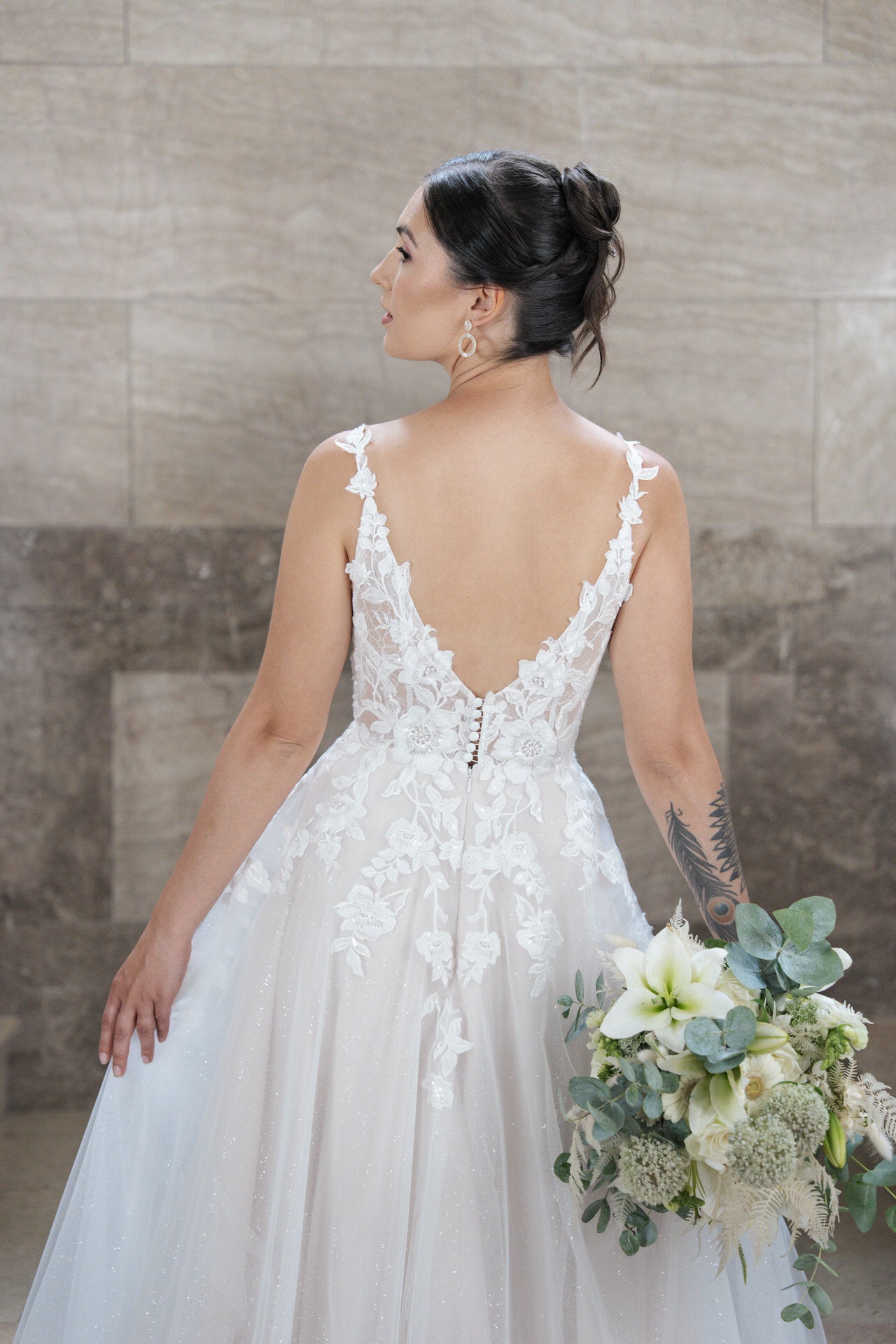 Eine Braut in einem weißen, mit Spitze besetzten Bridonista V26-A22 Hochzeitskleid steht mit dem Rücken zur Kamera und hält einen Strauß weißer Lilien und Grünpflanzen vor einem neutralen Steinmauer-Hintergrund.