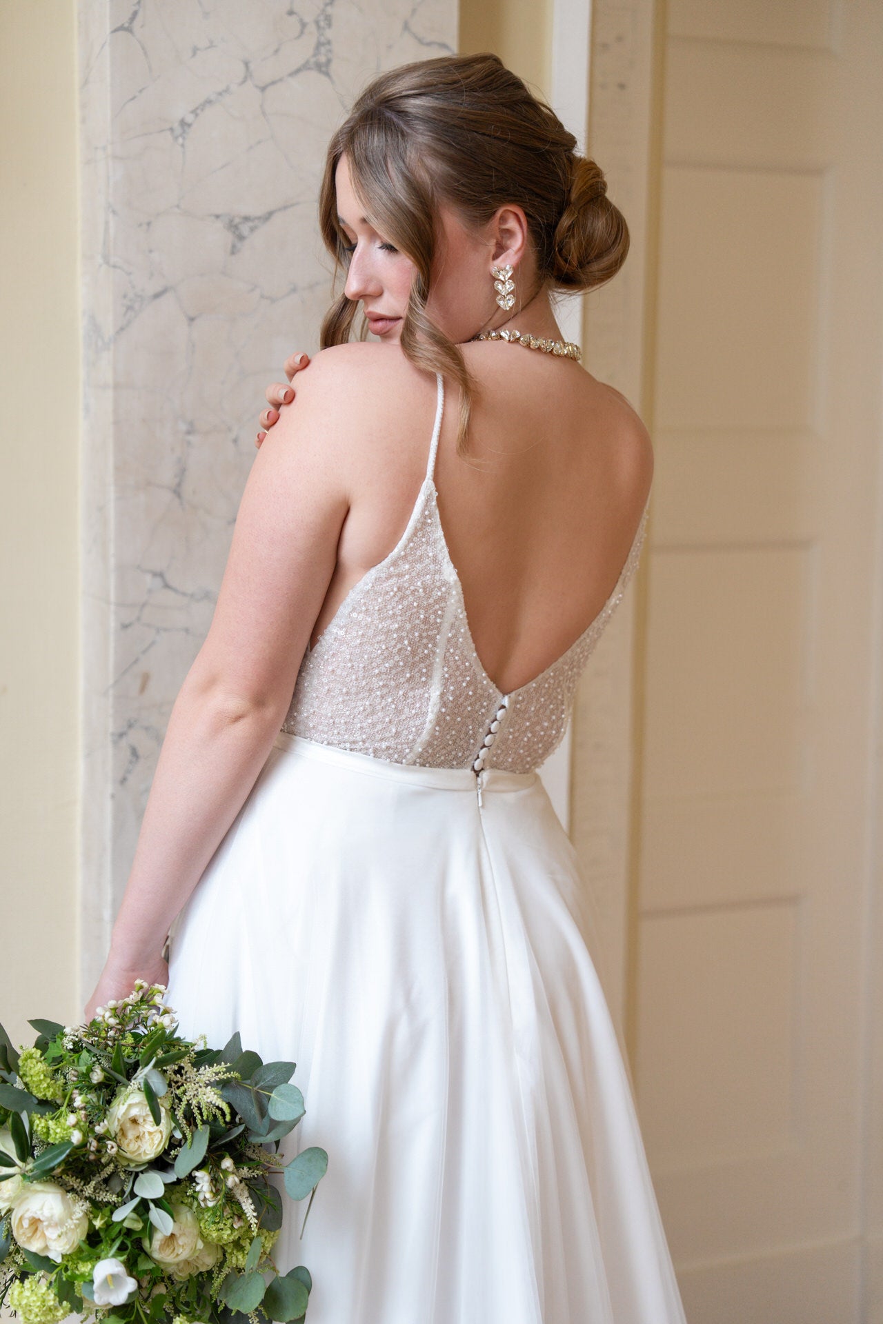 Eine Braut im weißen, ärmellosen Bridonista V26-A23 Brautkleid mit tiefem Rücken und Perlendetails hält einen grün-weißen Strauß und lächelt sanft über ihre Schulter. Ihre lockere Hochsteckfrisur bietet die perfekte Inspiration für Brautmoden.