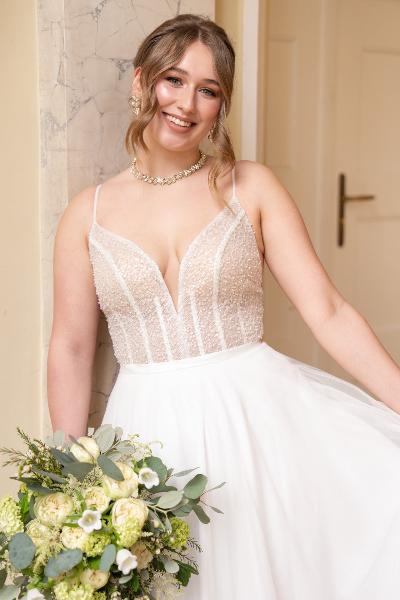 Eine lächelnde Braut im Bridonista V26-A23 Perlenbrautkleid hält einen Strauß weißer und grüner Blumen, während sie in einem Innenraum vor einer Marmorwand und einer Tür steht.