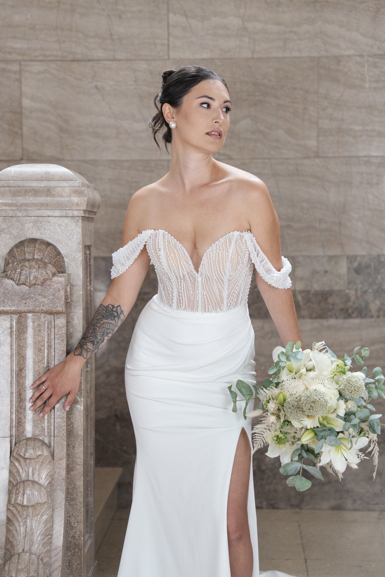 Eine Frau im schulterfreien weißen Brautkleid V26-F01 von Bridonista hält einen weiß-grünen Blumenstrauß in der Hand. Sie steht an einer Steinmauer, hat ihr Haar zu einer Hochsteckfrisur frisiert und blickt mit einem Unterarmtattoo zur Seite.