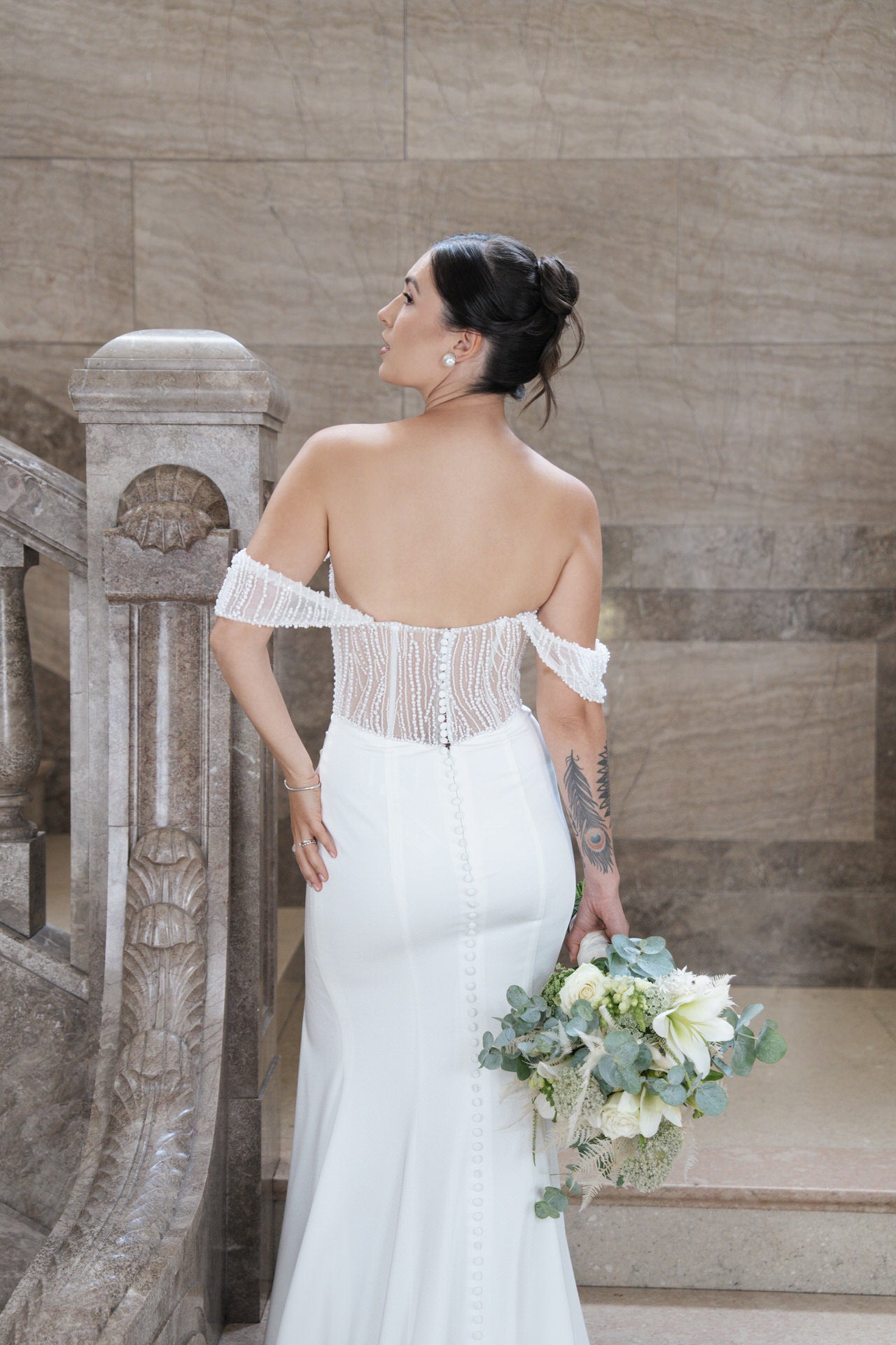 Eine Braut in dem schulterfreien weißen Kleid Bridonista V26-F01 steht auf einer Steintreppe und hält einen Strauß weißer und grüner Blumen. Sie blickt von der Kamera weg, ihr dunkles Haar ist zu einer eleganten Hochsteckfrisur frisiert.