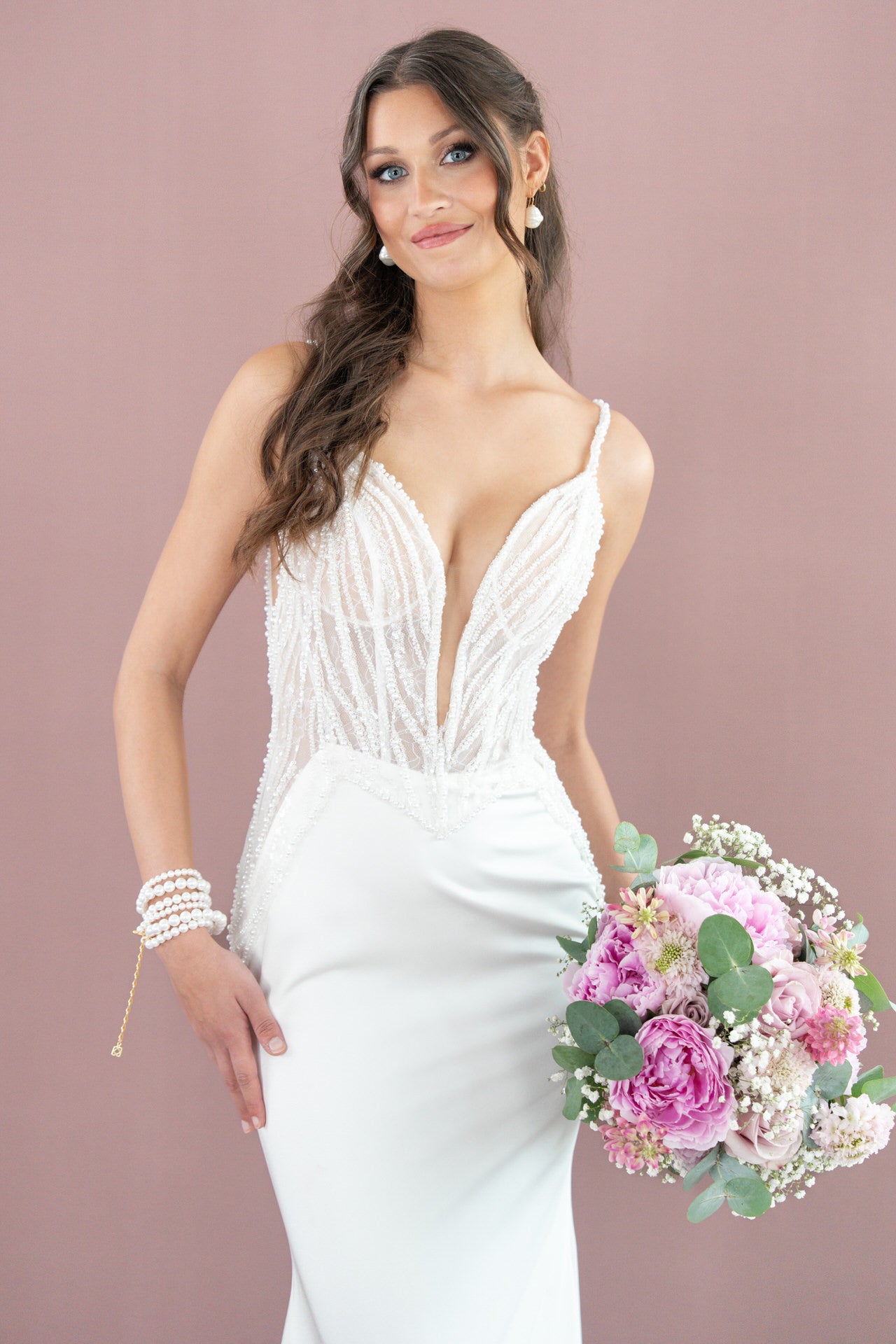 Eine Frau im weißen, perlenbesetzten Bridonista V26-F02 Brautkleid hält einen Strauß mit rosa und weißen Blumen. Mit langen, gewellten braunen Haaren und einem leichten Lächeln vor einem malvenfarbenen Hintergrund bietet sie die perfekte Brautmoden-Inspiration.