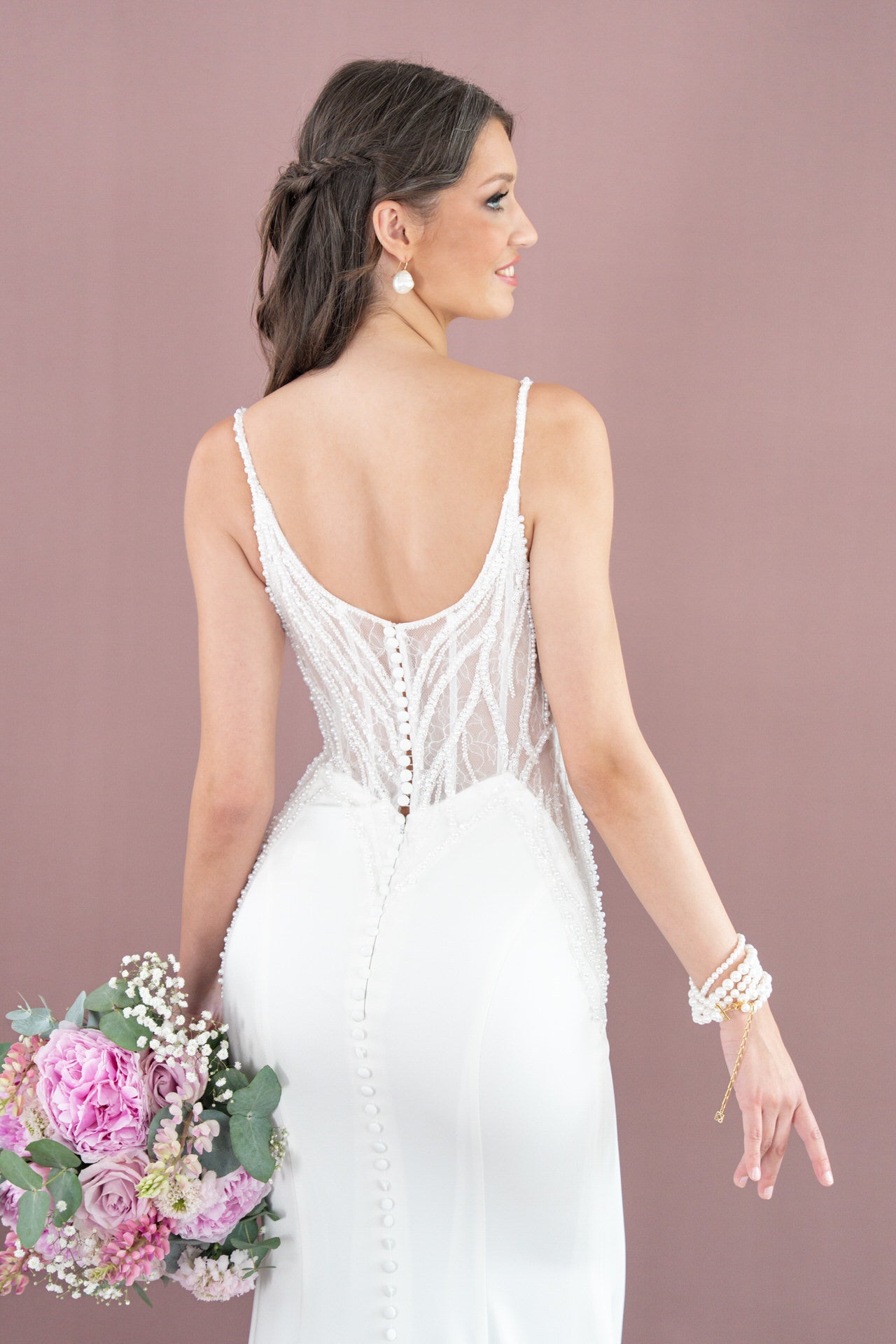 Eine Frau im perlenbesetzten weißen Brautkleid V26-F02 von Bridonista mit tiefem Rücken hält rosa und lila Blumen. Mit dem Gesicht nach außen zeigt sie Knöpfe und Perlenschmuck, während ein hellvioletter Hintergrund dieses elegante Hochzeitskleid hervorhebt.