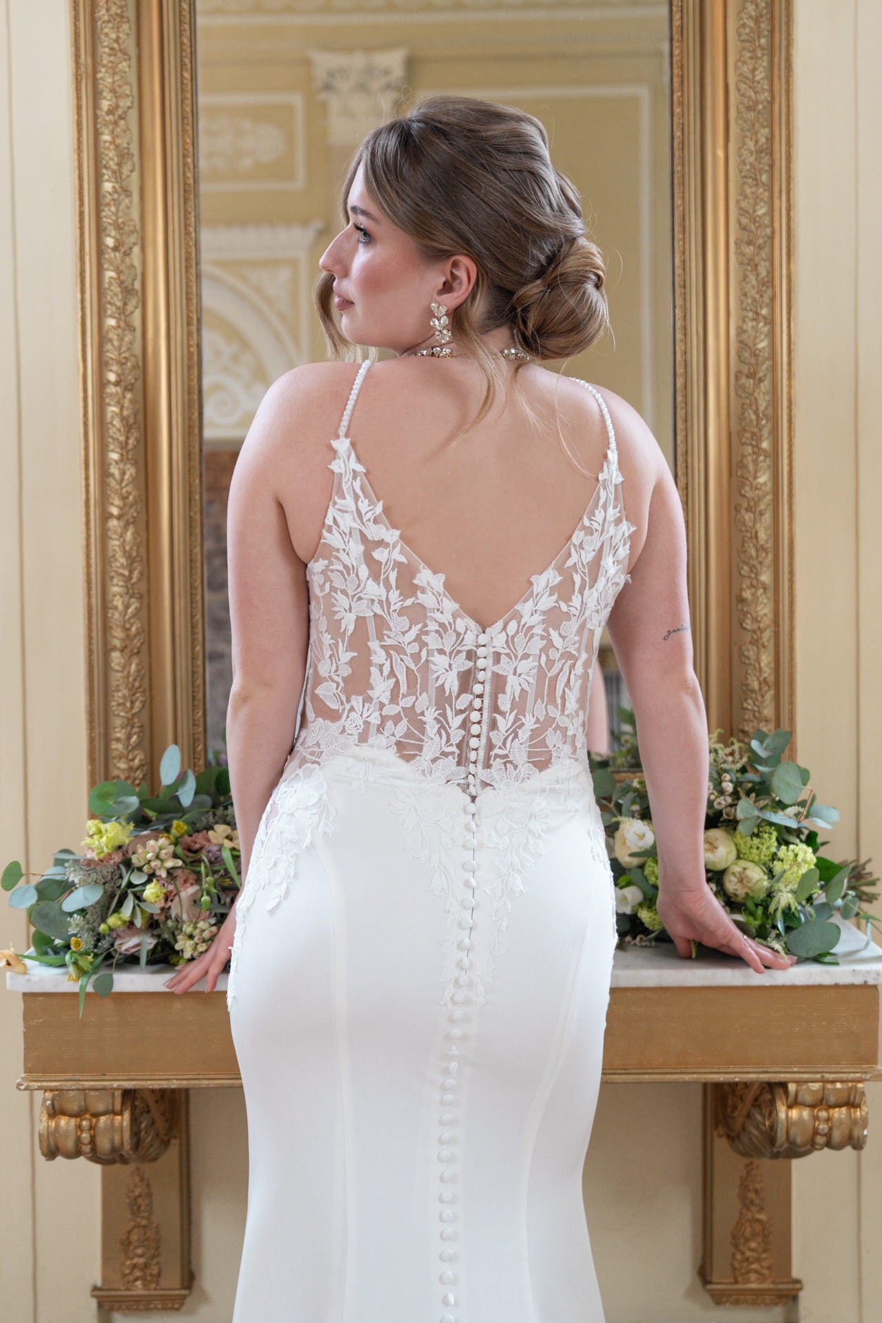 Eine Frau im Bridonista V26-F03 Brautkleid mit Spitzenrücken steht mit hochgestecktem Haar vor einem Spiegel und blickt zur Seite. Blumenarrangements vervollständigen die Szene - die perfekte Inspiration für Brautmoden.