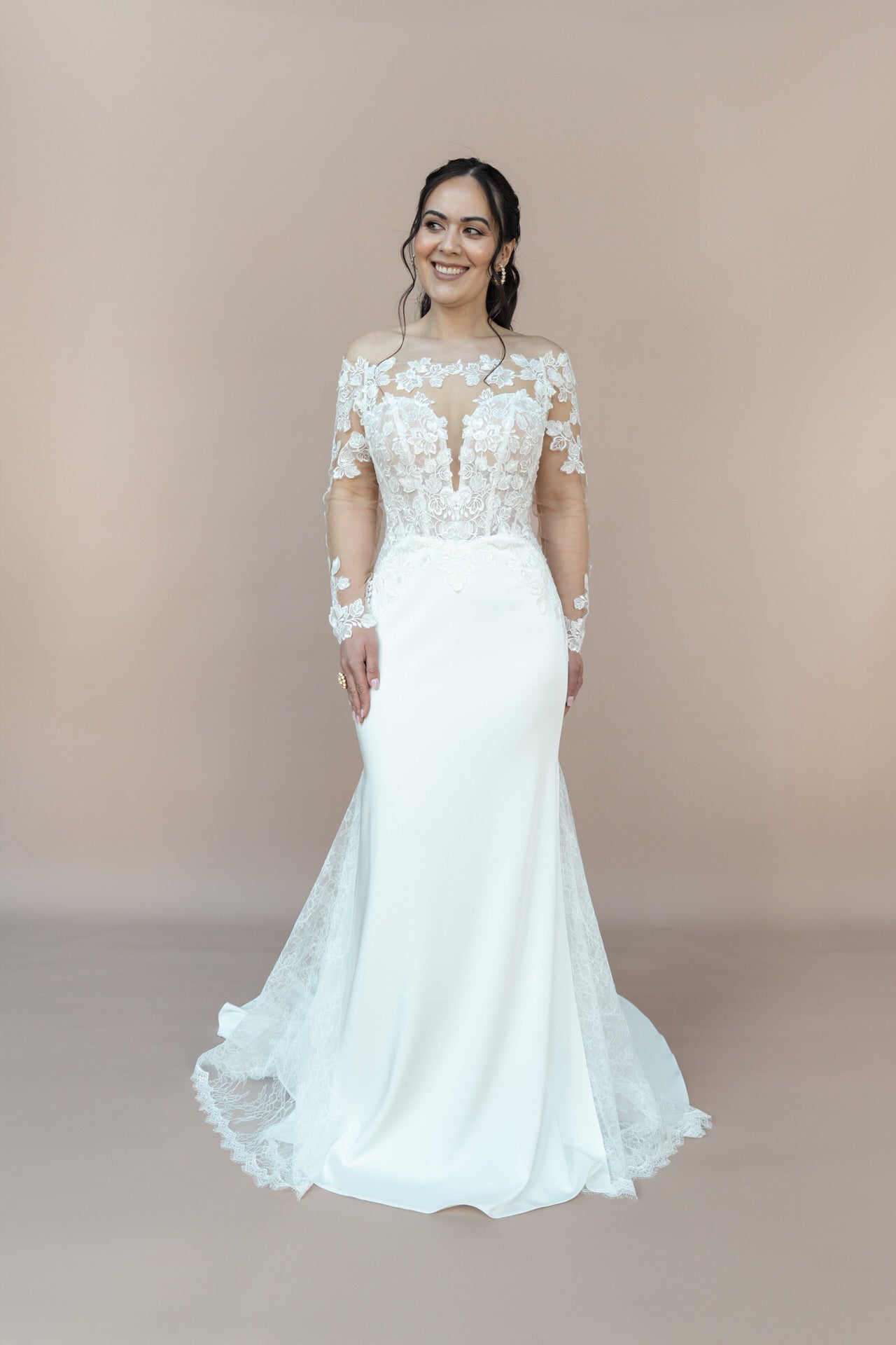 Eine Frau steht vor einem beigen Hintergrund, lächelt und blickt zur Seite, während sie das lange weiße Brautkleid Bridonista V26-F05 mit Spitzenärmeln und Blumenstickerei trägt.