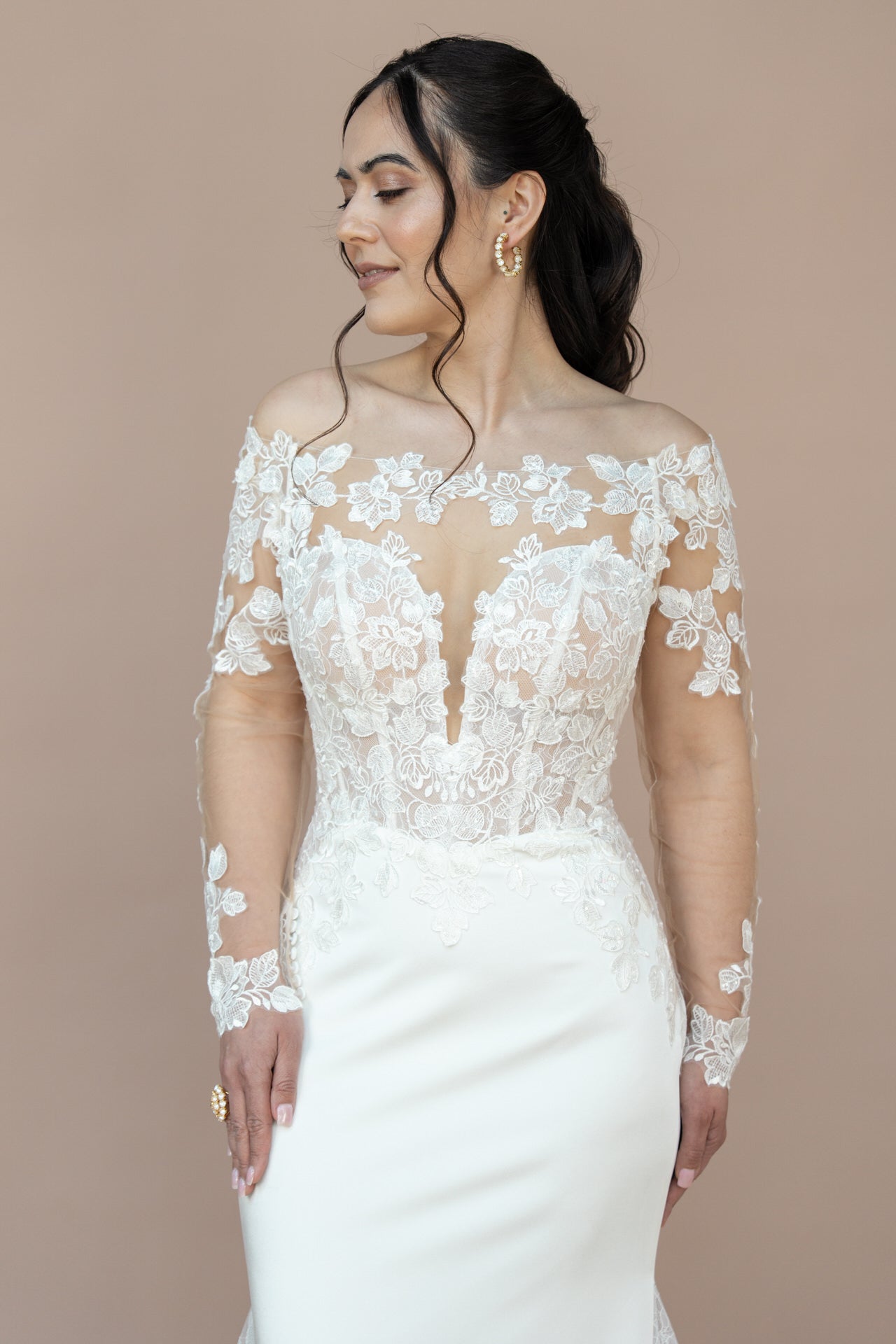 Eine Frau in dem schulterfreien, langärmeligen weißen Brautkleid Bridonista V26-F05 mit floraler Spitze steht vor einem beigen Hintergrund und blickt mit einem sanften Lächeln zur Seite - zeitlose Brautmoden-Eleganz.