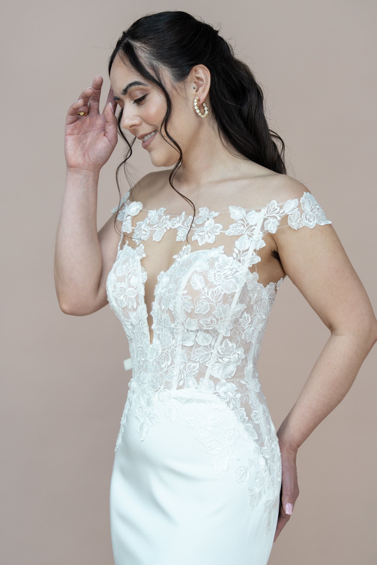 Eine Frau in einem weißen schulterfreien Bridonista V26-F05-Brautkleid aus Spitze lächelt und berührt ihr Gesicht. Sie hat langes, gewelltes Haar und goldene Ohrringe und steht vor einem schlichten hellbraunen Hintergrund.