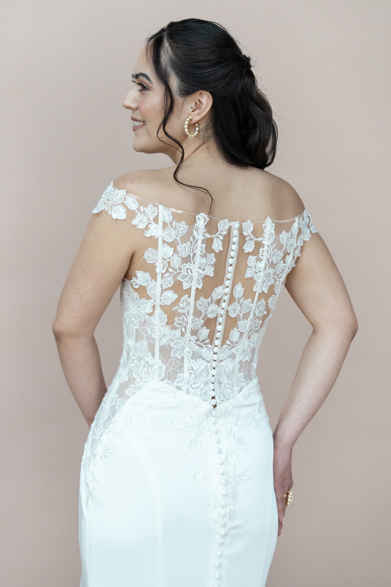 Eine Frau trägt das Bridonista V26-F05, ein weißes, schulterfreies Brautkleid mit floralen Spitzendetails am Rücken. Sie schaut lächelnd über die Schulter und trägt dunkle, lockere Wellen und goldene Ohrringe.