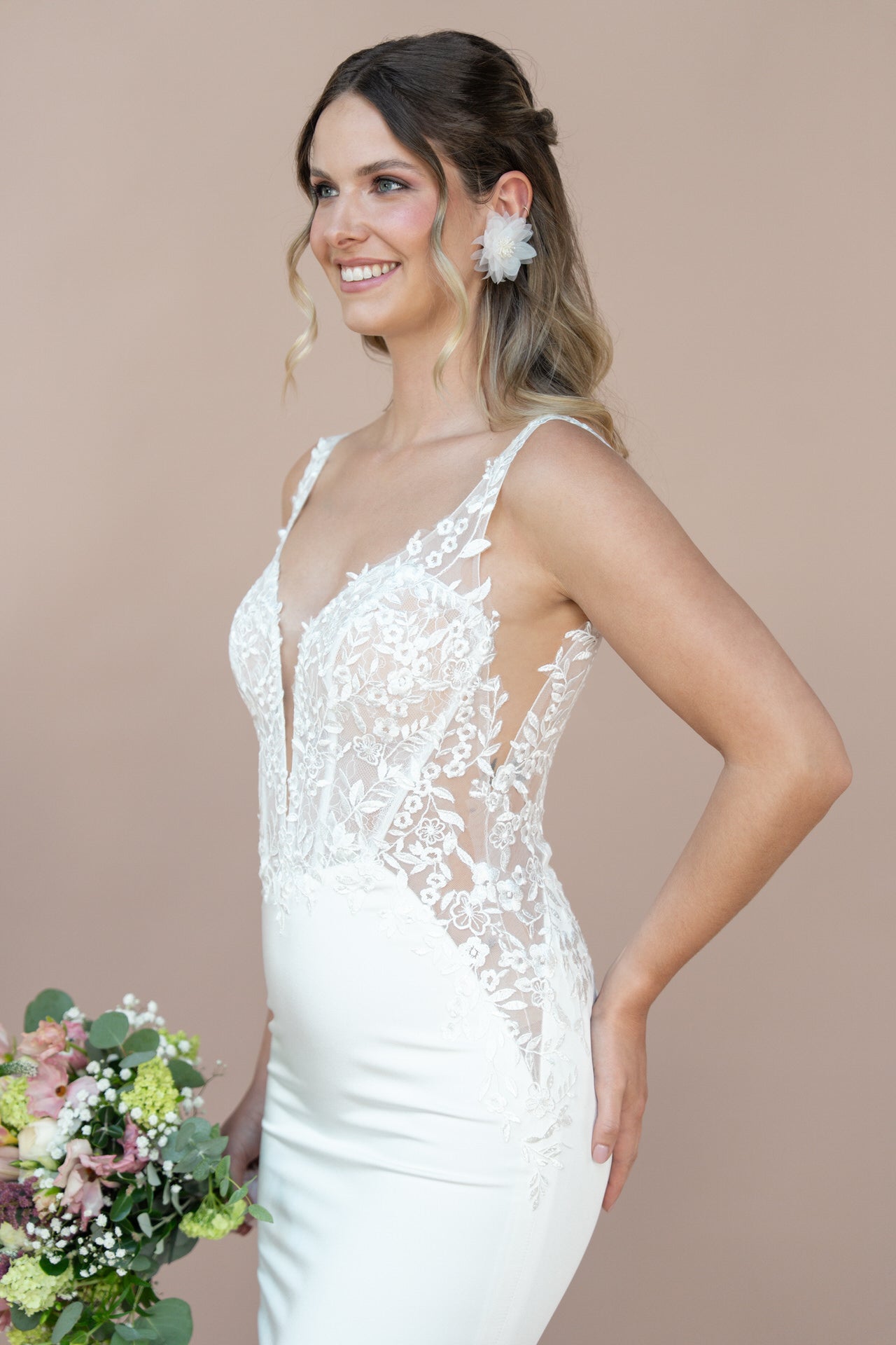 Eine Frau im weißen, spitzenverzierten Bridonista V26-F07 Brautkleid hält einen Blumenstrauß, lächelt mit gewelltem Haar und einem floralen Accessoire vor einem schlichten beigen Hintergrund - die perfekte Inspiration für Brautmoden.