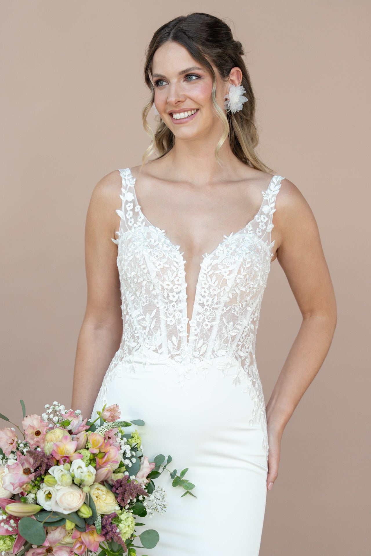 Eine lächelnde Frau im weißen Hochzeitskleid Bridonista V26-F07 hält einen Strauß mit rosa, gelben und weißen Blumen vor einem schlichten beigen Hintergrund. Sie hat gewelltes Haar und trägt einen großen geblümten Ohrring.