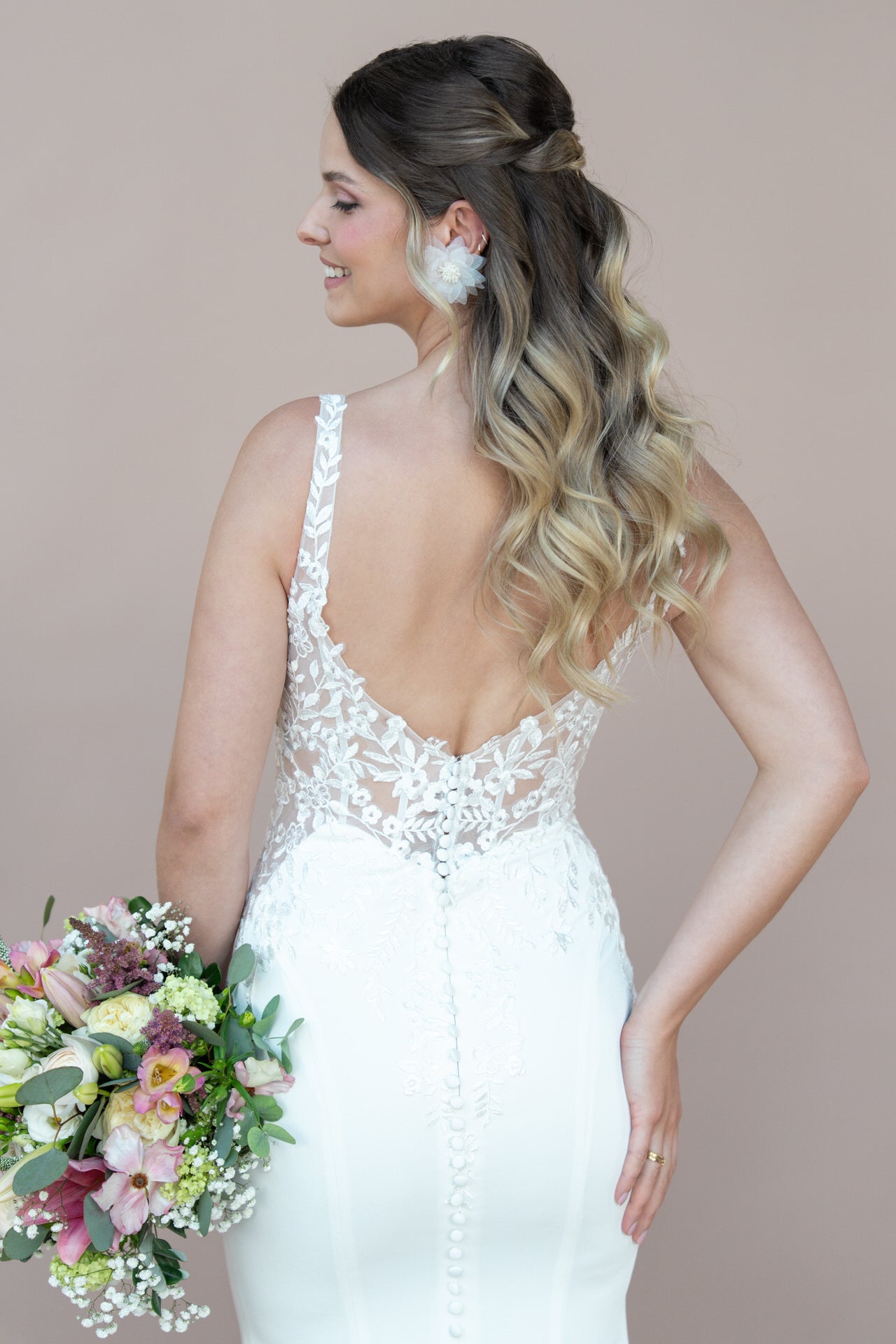 Eine Braut mit gewelltem, ombréfarbenem Haar hält einen Blumenstrauß und trägt das Bridonista V26-F07, ein elegantes, modernes weißes Spitzen-Hochzeitskleid mit tiefem Rücken und Zierknöpfen. Sie wendet sich ab und lächelt über ihre Schulter.