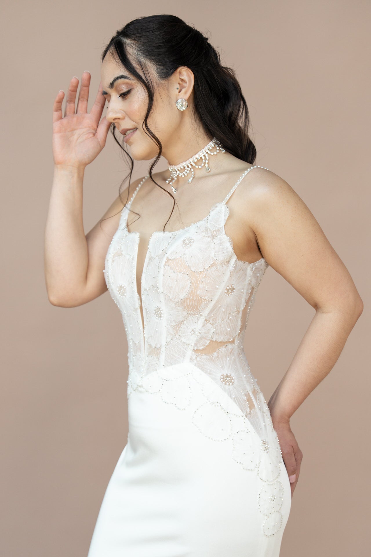 In Bridonista V26-F08 - einem weißen, ärmellosen Brautkleid mit floraler Spitze - posiert diese Frau vor einem beigen Hintergrund und lächelt sanft mit lockeren Wellen in ihrem dunklen Haar, perfekt für die Brautmoden-Inspiration.