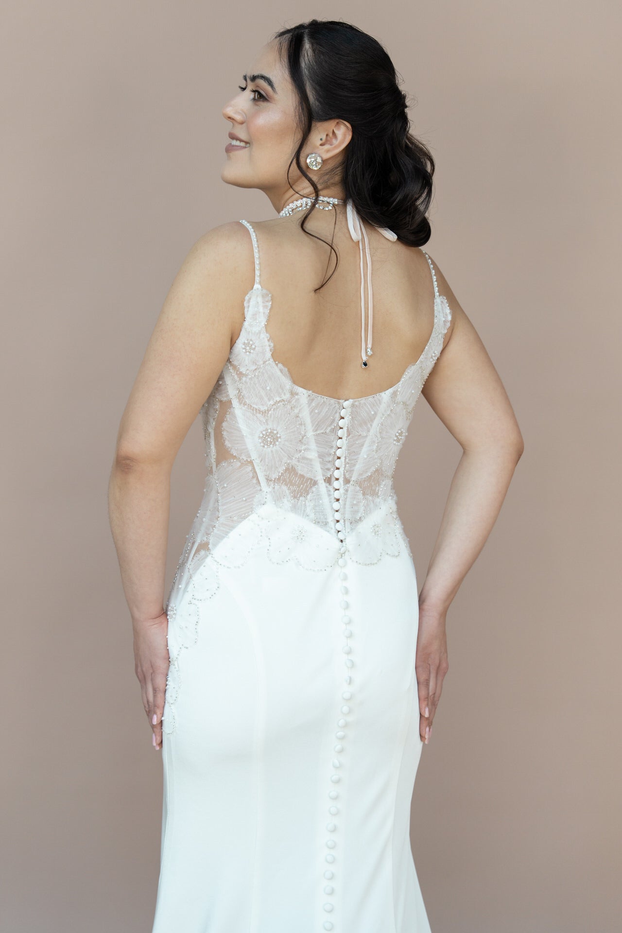 Eine Frau trägt das elegante Brautkleid Bridonista V26-F08 mit feinen Spitzendetails, Knopfleiste und offenem Rücken. Ihr dunkles Haar ist zu einem tiefen Updo gestylt und sie lächelt zur Kamera.