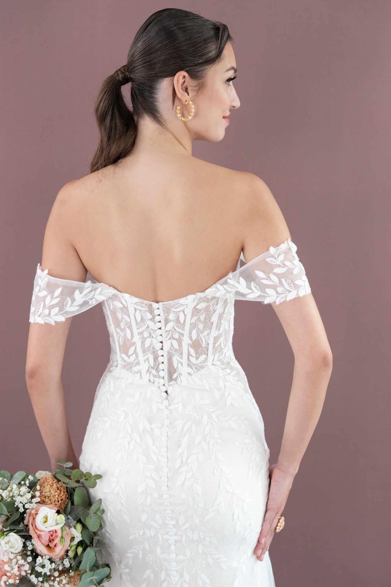 Frau in Bridonista V26-F09, einem schulterfreien Spitzenbrautkleid mit floralen Details, mit einem Blumenstrauß in der Hand, von hinten gesehen vor einem lilafarbenen Hintergrund - eine ideale Inspiration für moderne Brautmode.