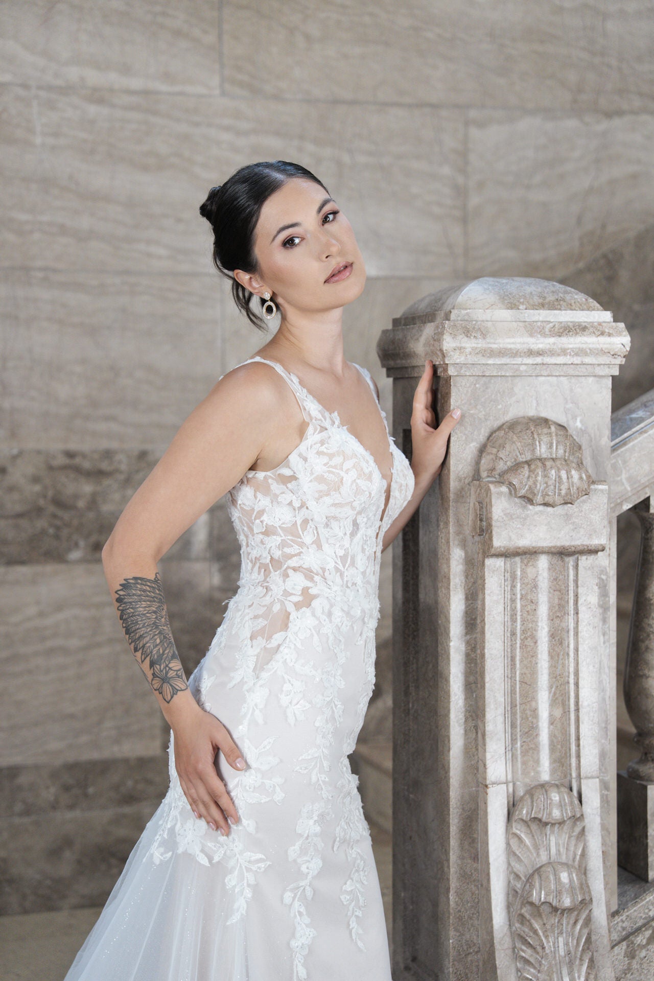 Eine Frau mit dunklem Haar in einer Hochsteckfrisur posiert vor einer Steintreppe und trägt das Bridonista V26-F12, ein weißes Spitzenbrautkleid mit Blumendetails. Sie trägt große Ohrringe und zeigt eine Tätowierung auf ihrem linken Unterarm.