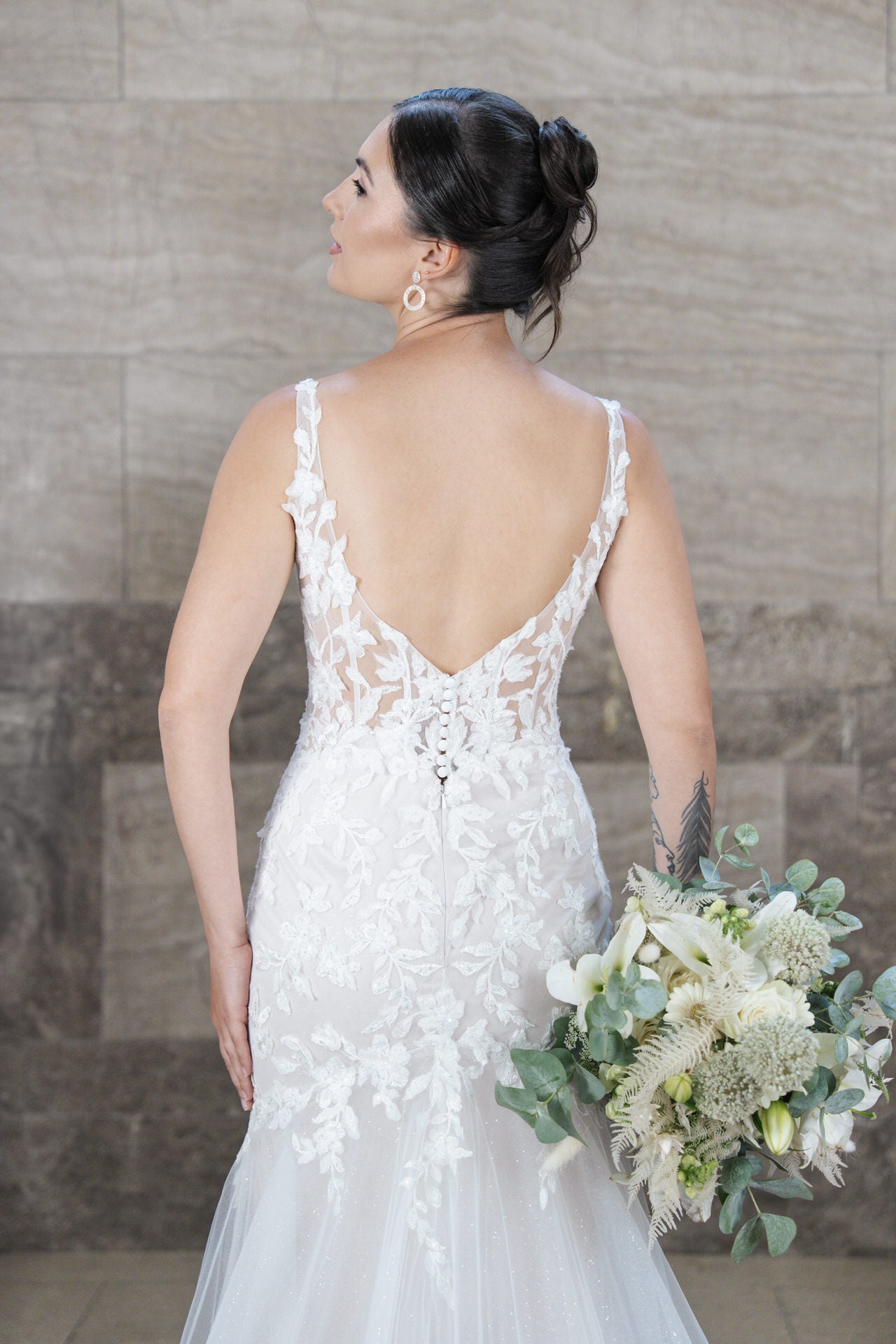 Eine Braut im weißen Spitzenhochzeitskleid Bridonista V26-F12 steht mit dem Rücken zur Kamera und hält einen Strauß aus weißen Blumen und Grünzeug. Ihr dunkles Haar ist zu einem Dutt gestylt, während sie anmutig vor einer Steinmauer posiert.