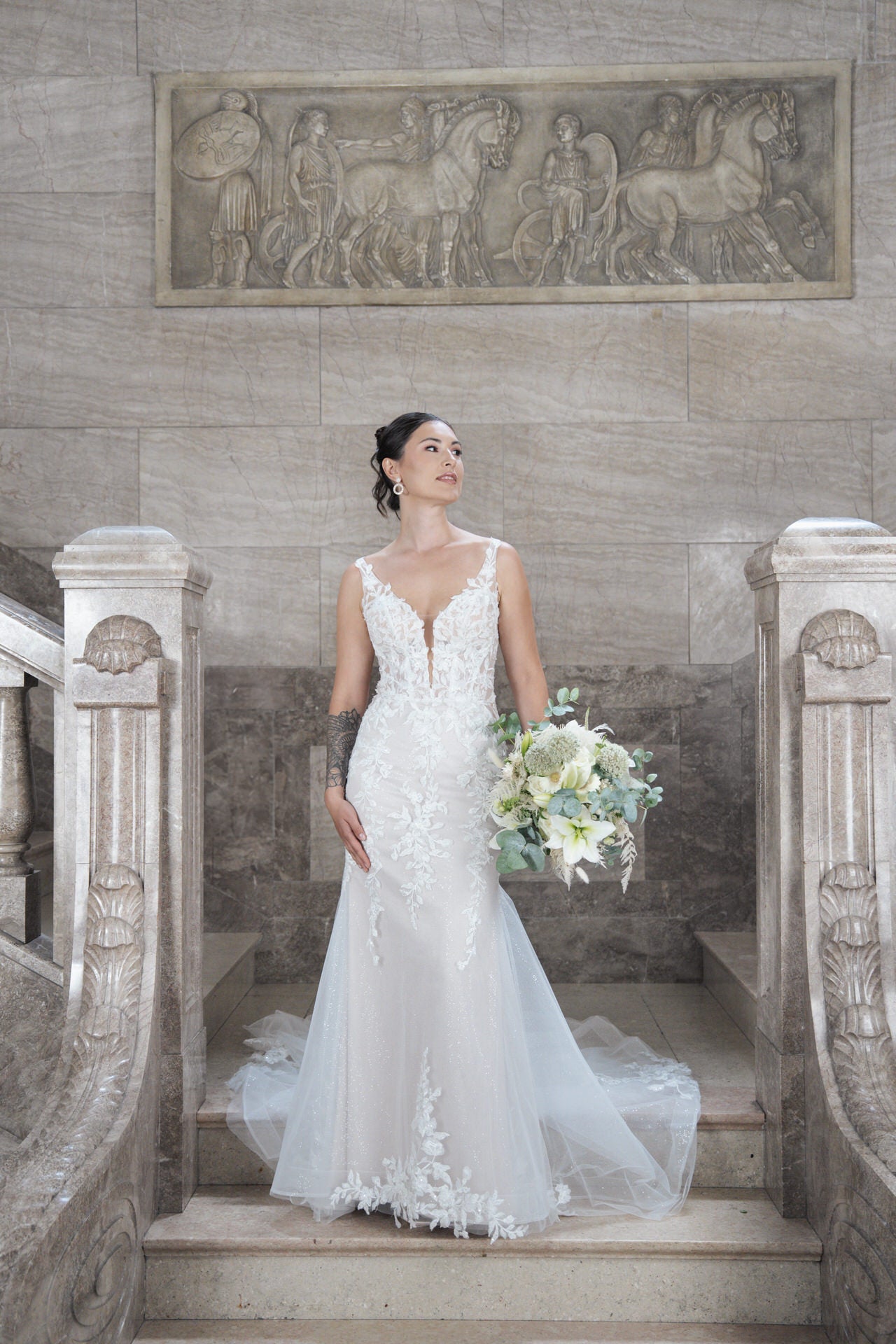 Eine Braut trägt das elegante Brautkleid Bridonista V26-F12 und steht mit Blumenstrauß auf Marmortreppen. Sie blickt nach oben, kunstvolle Reliefs schmücken die Wand-perfektes Ambiente für stilvolle Brautmode.