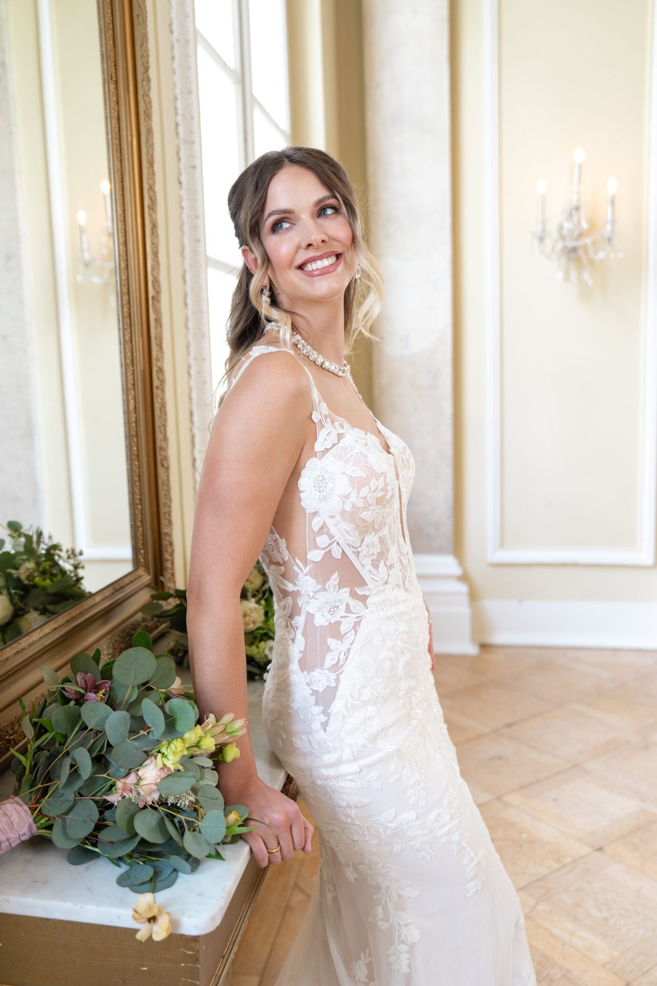 Eine lächelnde Frau im ärmellosen Brautkleid aus Spitze von Bridonista V26-F13 steht in einem Innenraum vor einem Spiegel und einem Blumenstrauß, während das Sonnenlicht durch ein Fenster mit eleganter Dekoration im Hintergrund fällt.