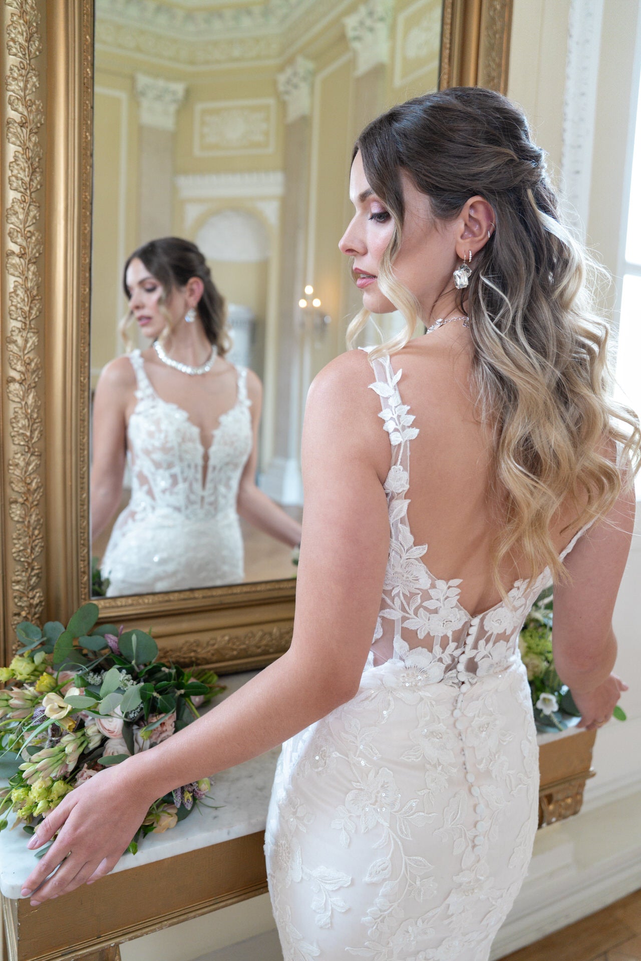 Eine Braut mit gewelltem Ombre-Haar steht vor einem Spiegel in einem geschmückten Raum. Sie trägt das Bridonista V26-F13 Spitzen-Hochzeitskleid und hält einen Strauß grüner und weißer Blumen in der Hand; ihr Spiegelbild ist inmitten des klassischen Dekors zu sehen.