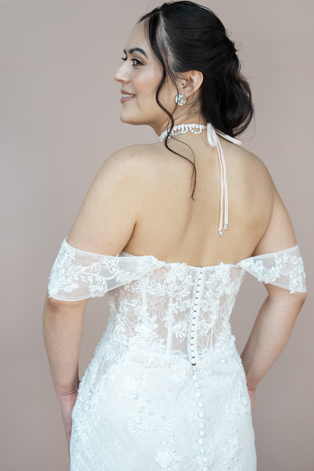 In einem Bridonista V26-M01, einem weißen Spitzen-Brautkleid im Off-Shoulder-Stil, steht eine Frau seitlich und lächelt. Ihr dunkles Haar ist elegant hochgesteckt, dazu trägt sie Perlenohrringe und eine Schleifen-Halskette.