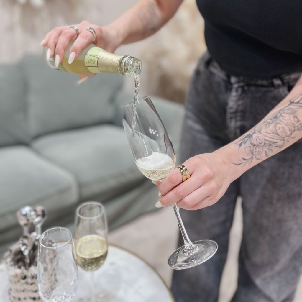 Eine Person mit tätowierten Armen schenkt Sekt in ein Glas ein. Zwei weitere gefüllte Gläser und ein dekorativer Gegenstand stehen auf einem Tisch vor einem grünen Sofa. Die Person trägt eine dunkle Jeans und ein schwarzes Oberteil.