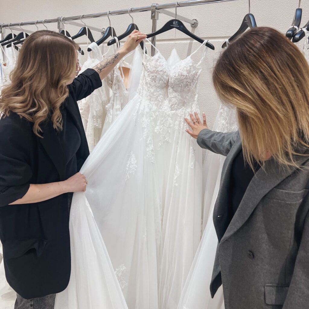 Zwei Frauen begutachten ein weißes, spitzenbesetztes Hochzeitskleid auf einem Kleiderbügel in einem Brautmodengeschäft, umgeben von anderen Brautkleidern.